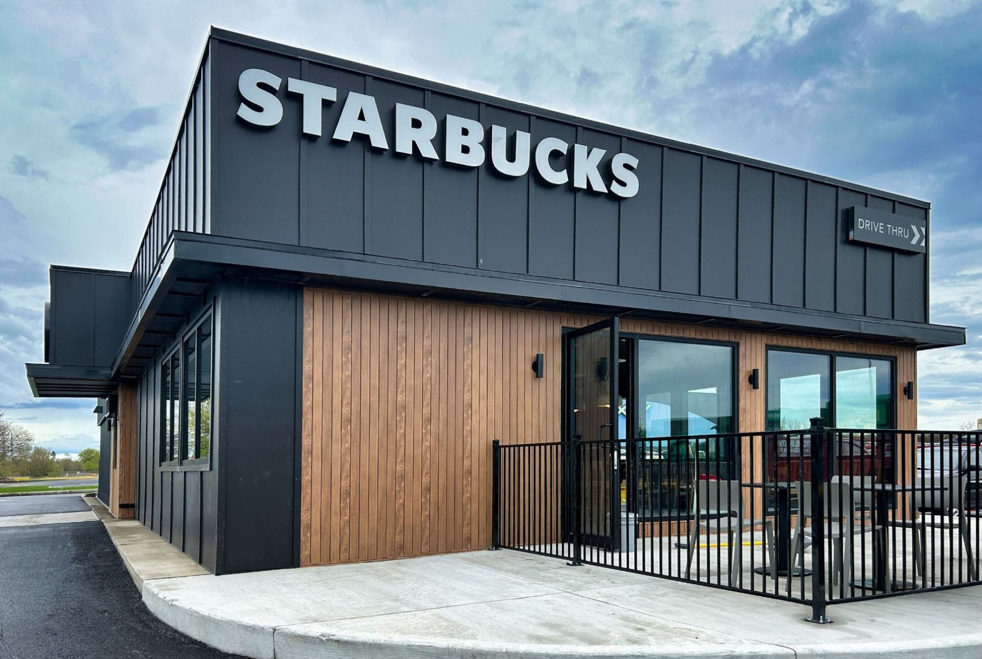 A modern Starbucks building with dark exterior panels, wood accents, and large windows. The white “STARBUCKS” sign is mounted above the entrance, with a drive‑thru sign and outdoor seating enclosed by a metal railing.
