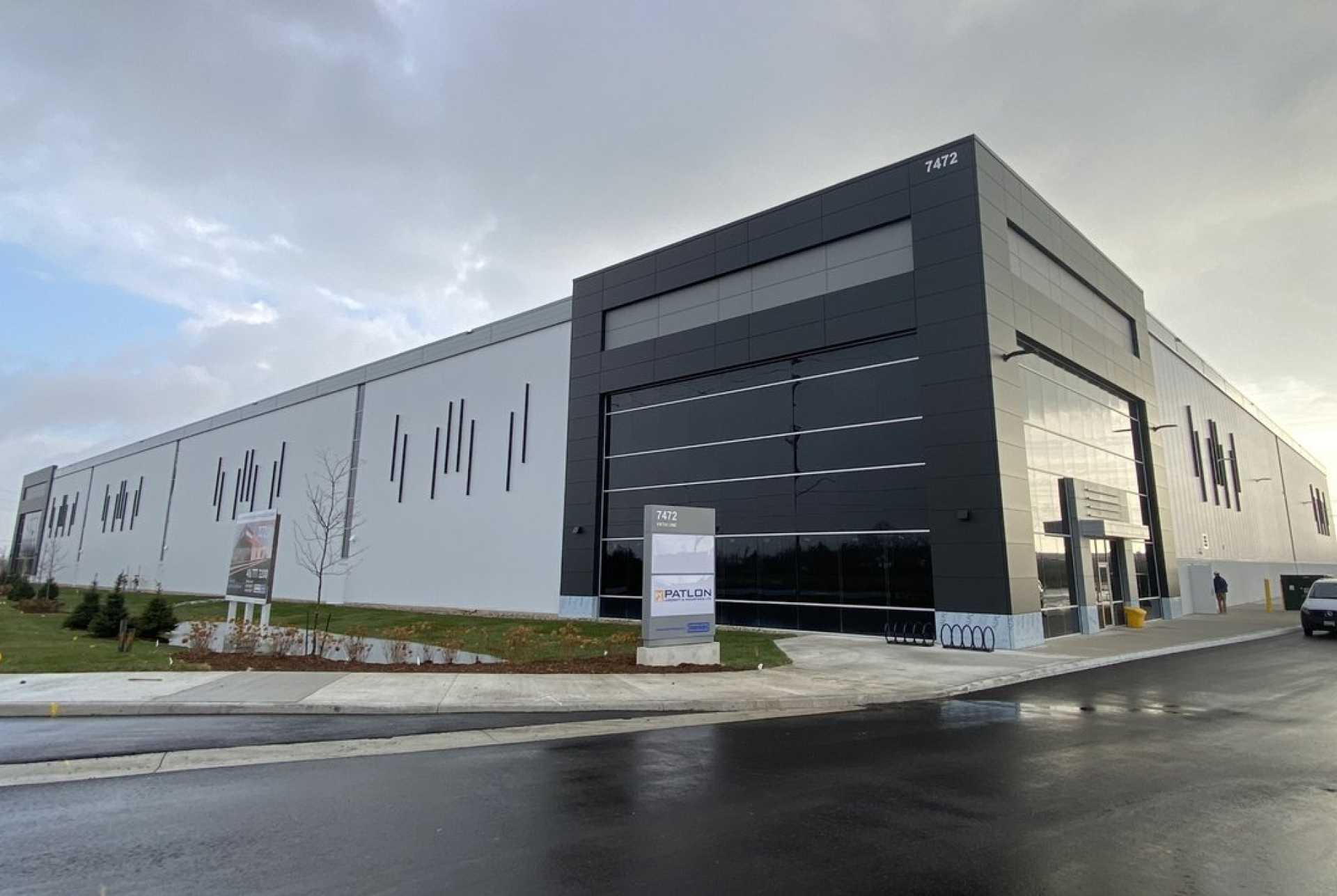 A modern industrial building with a white and dark gray exterior features clean horizontal lines and large glass panels. The structure sits along a paved roadway with landscaped grass, signage near the entrance, and an overcast sky.