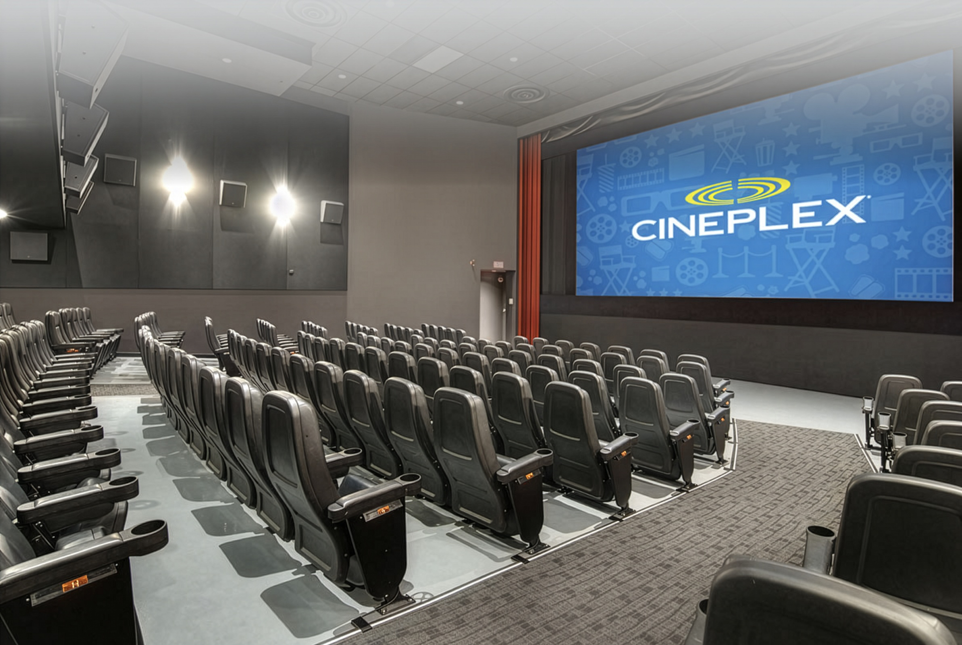 A Cineplex movie theatre auditorium with rows of black reclining seats facing a large screen displaying the Cineplex logo. Wall lights, tiered seating, and wide aisles create a spacious, modern cinema setting.