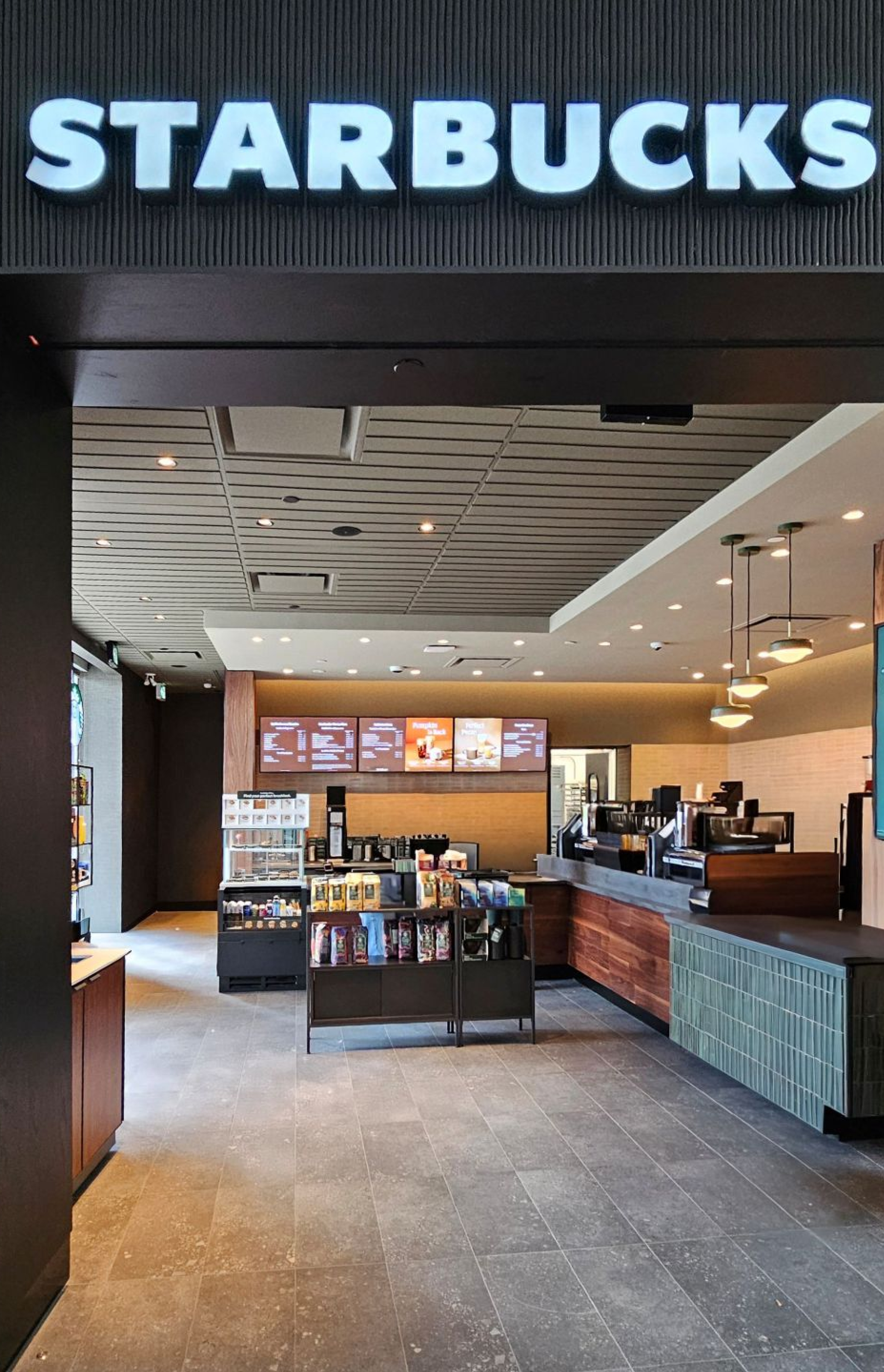 A Starbucks café interior featuring a wood‑accented service counter, menu boards, coffee equipment, and retail displays. Neutral tile floors, pendant lights, and an open layout create a clean, modern setting.