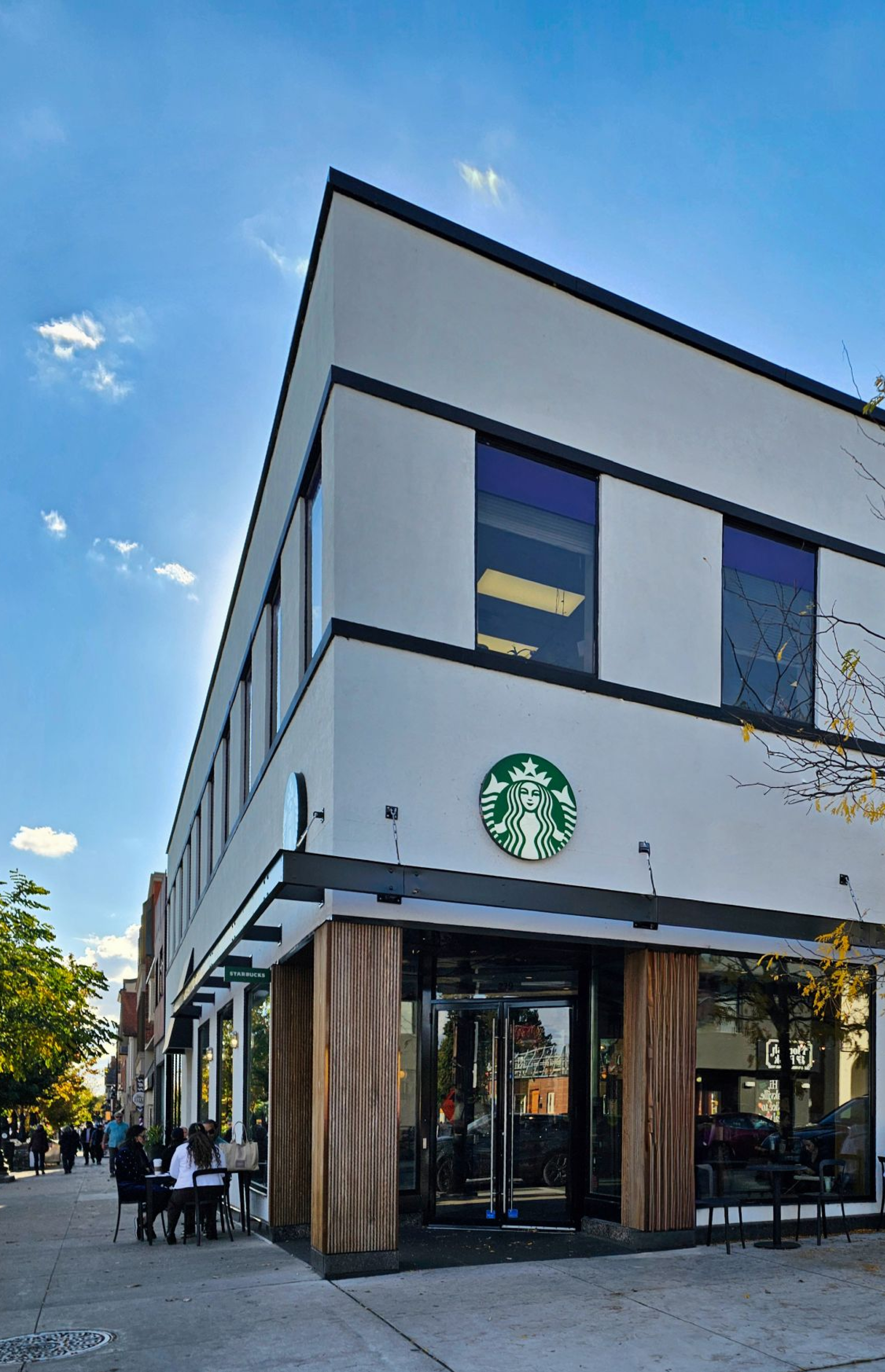 A modern corner Starbucks in a two‑storey building with a light‑coloured exterior, large windows, and wood column accents at the entrance. The green Starbucks logo is mounted above the glass doors along a city sidewalk.