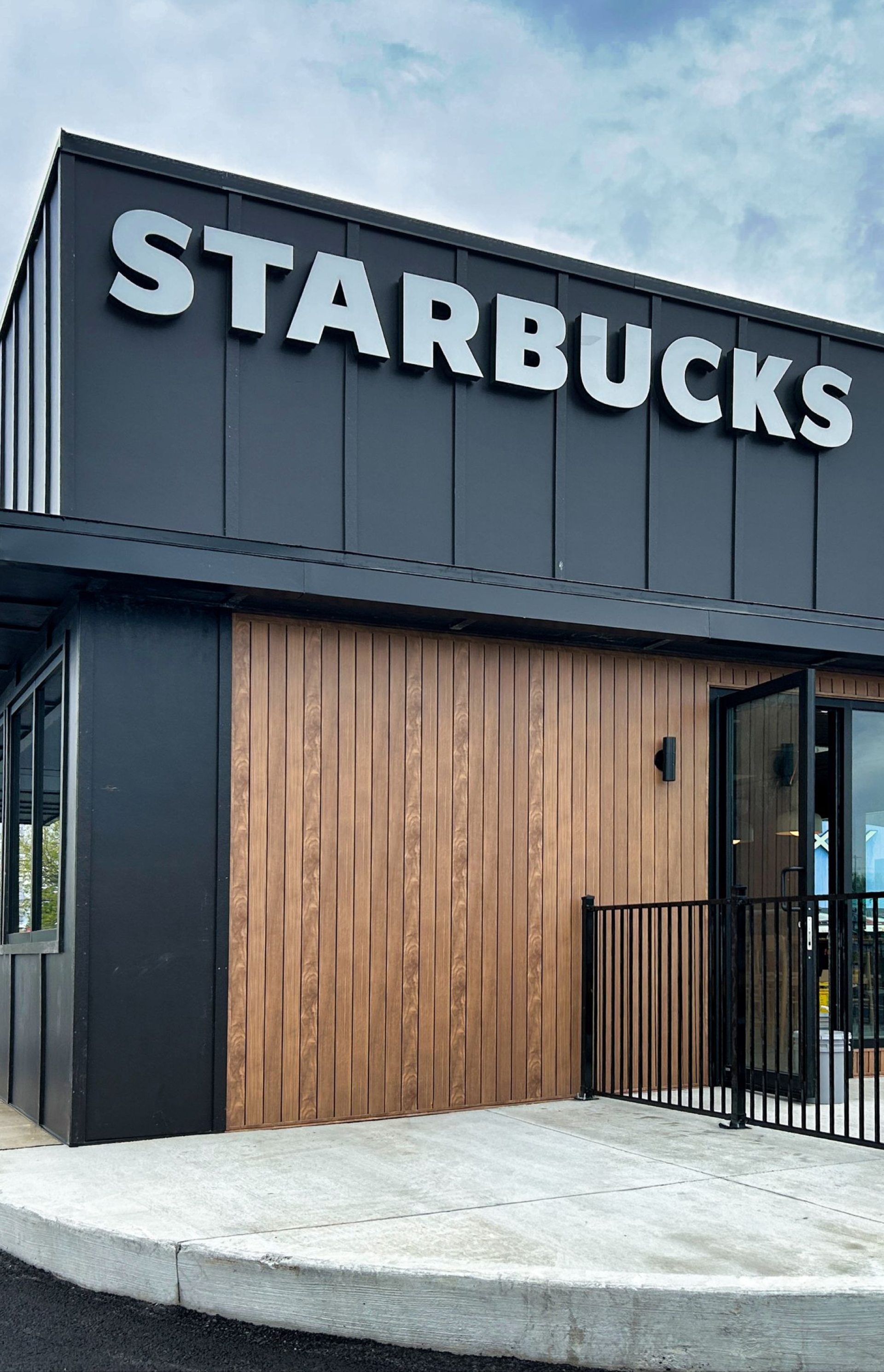 A modern Starbucks building with a dark metal exterior, wood accents, and large windows. The white “STARBUCKS” sign is mounted above the entrance, with an outdoor seating area enclosed by a black metal railing.