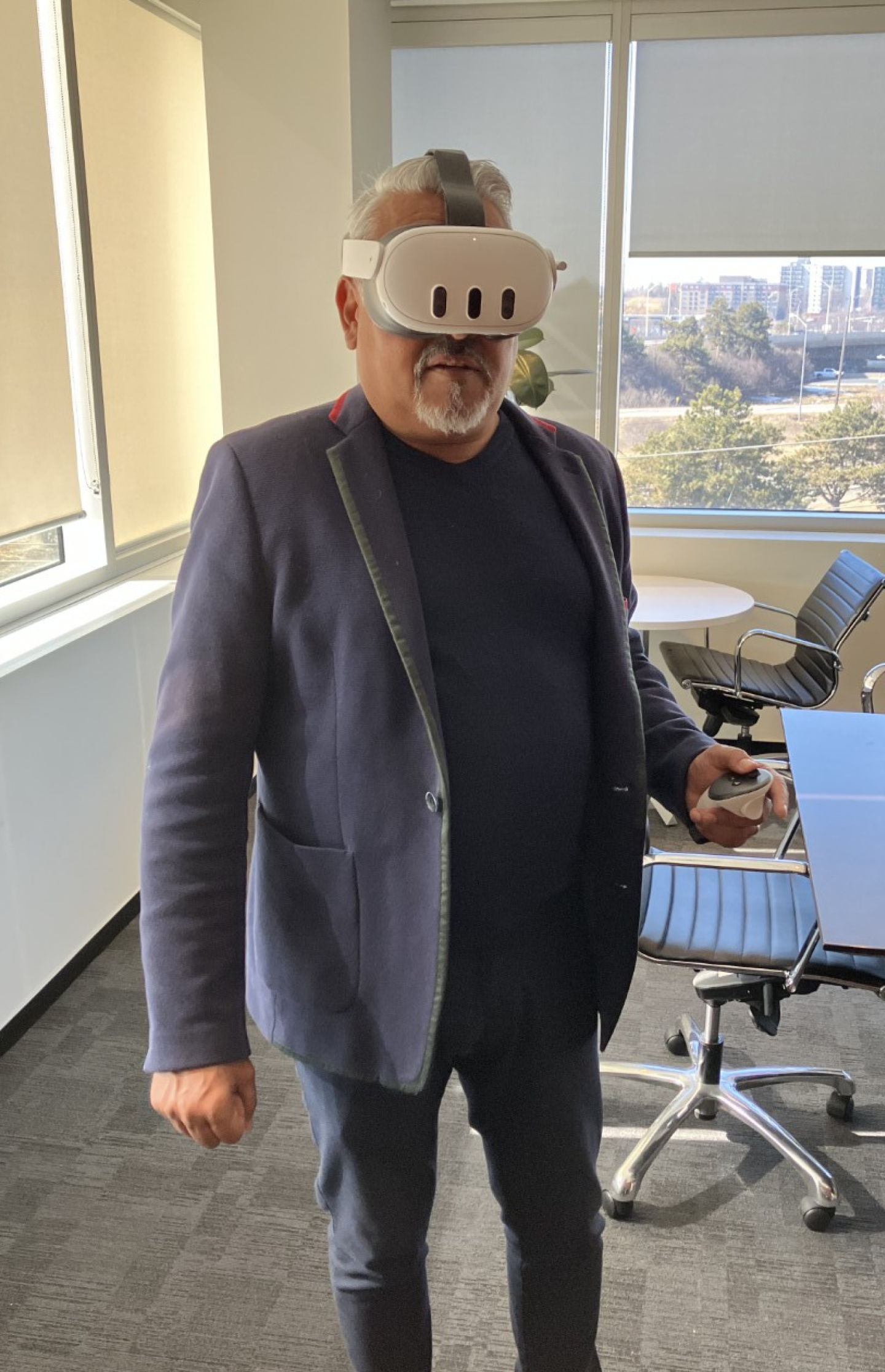 An office meeting room where a person stands wearing a virtual reality headset and holding a handheld controller. Tables, chairs, large windows with blinds, and a city view are visible in the background.
