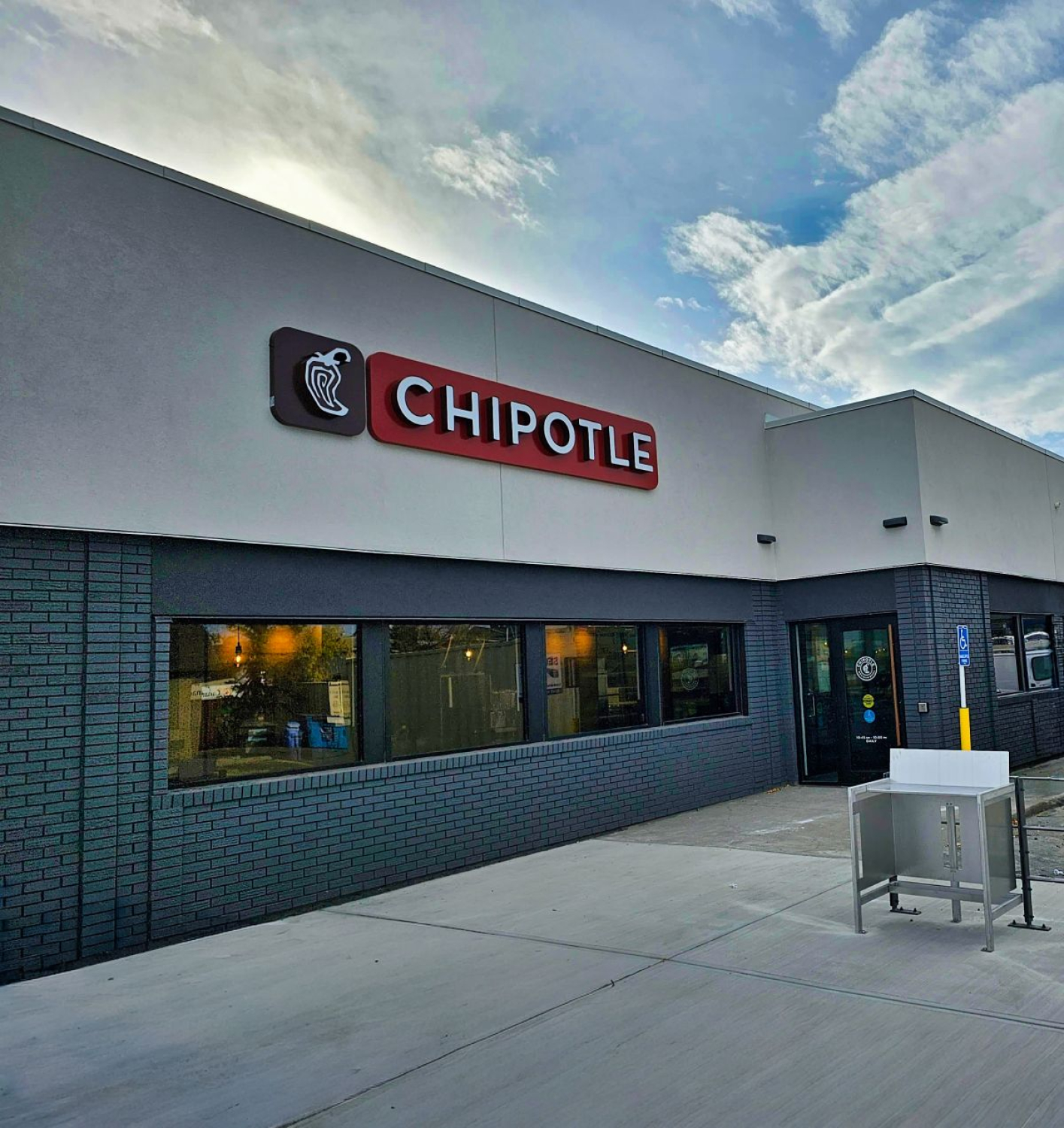 A Chipotle restaurant exterior with a light gray facade, dark brick walls, and large windows. The red “CHIPOTLE” sign and logo are mounted above the entrance, with a sidewalk and metal table visible outside.
