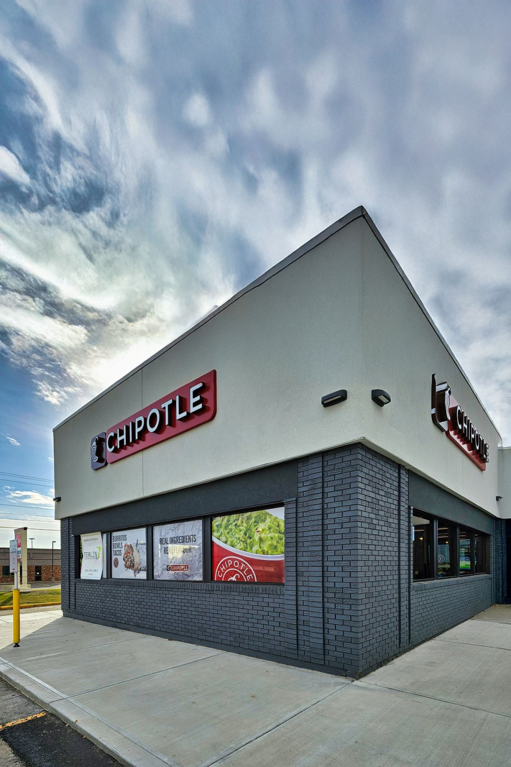 A Chipotle restaurant exterior with a light gray upper facade, dark brick base, and large windows wrapping a corner of the building. Red Chipotle signs are mounted on both visible sides beneath a partly cloudy sky.