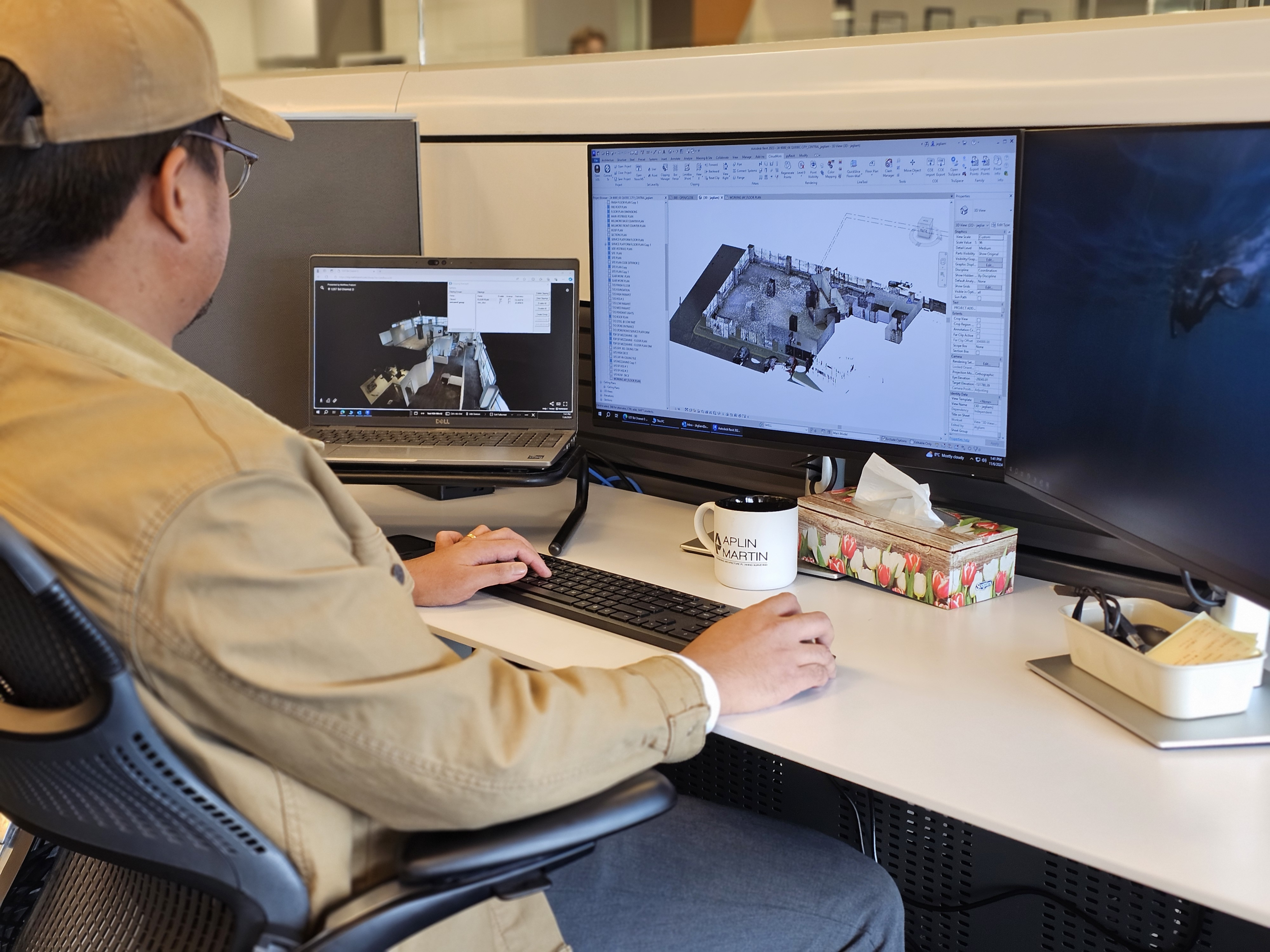 A person sits at an office workstation reviewing a 3D scan and building model on a laptop and a large monitor. The desk includes a keyboard, coffee mug, and tissue box within a modern office setting.