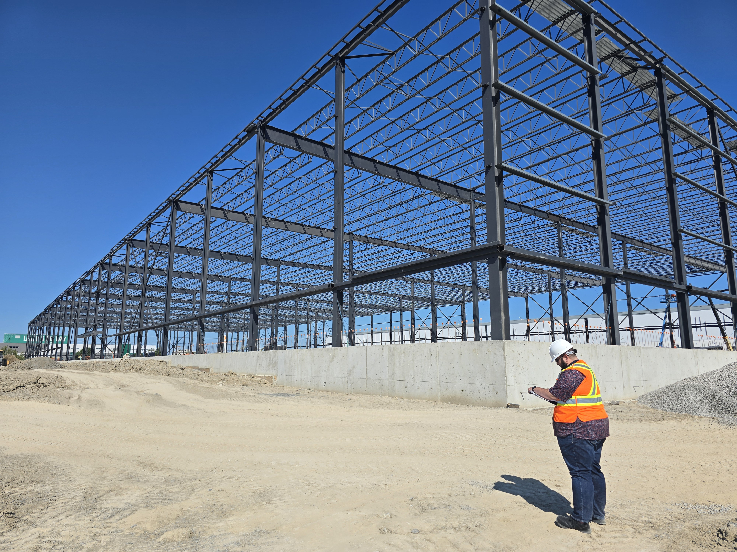 An industrial construction site showing a large steel building frame under assembly on a concrete foundation. A worker in safety gear stands on the dirt ground nearby, reviewing plans beneath a clear blue sky.