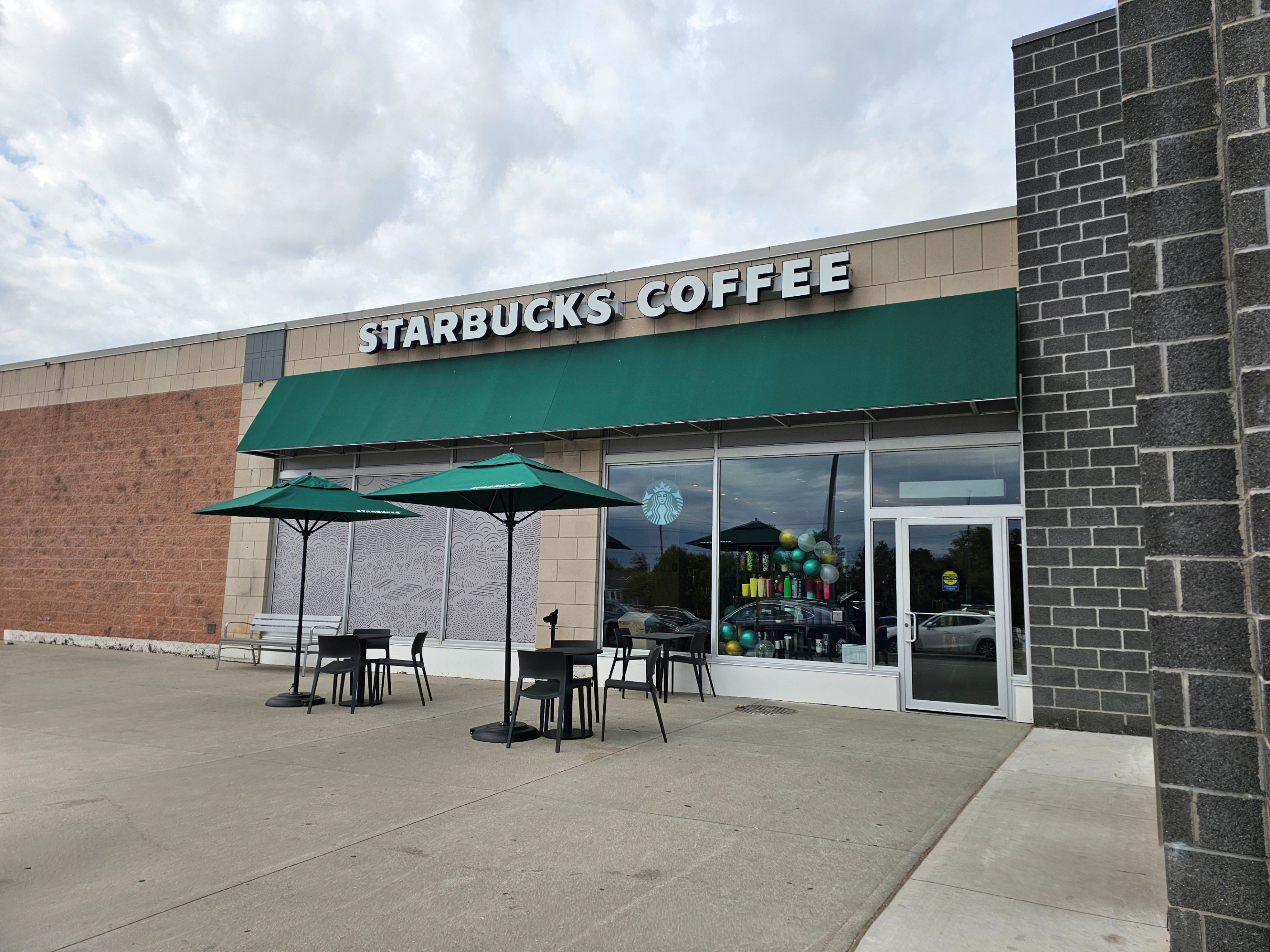 A Starbucks Coffee storefront with a beige and brick exterior, green awning, and large glass windows. Outdoor tables with green umbrellas sit on a wide concrete patio in front of the entrance under a cloudy sky.