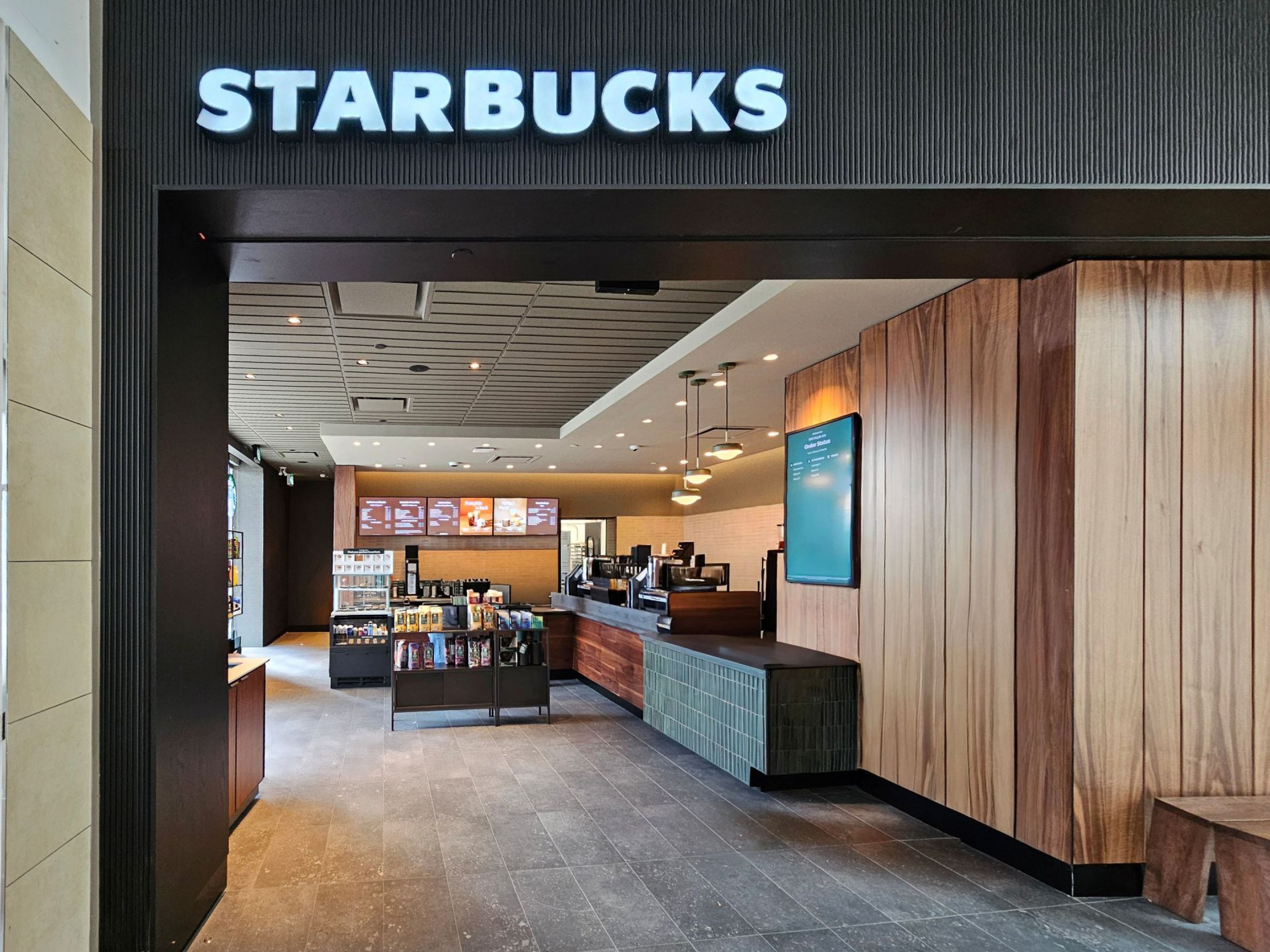 A Starbucks store entrance viewed from inside, showing a wood‑paneled service counter, menu boards, and retail displays. The illuminated “STARBUCKS” sign frames the opening into the café’s modern, warmly lit interior.
