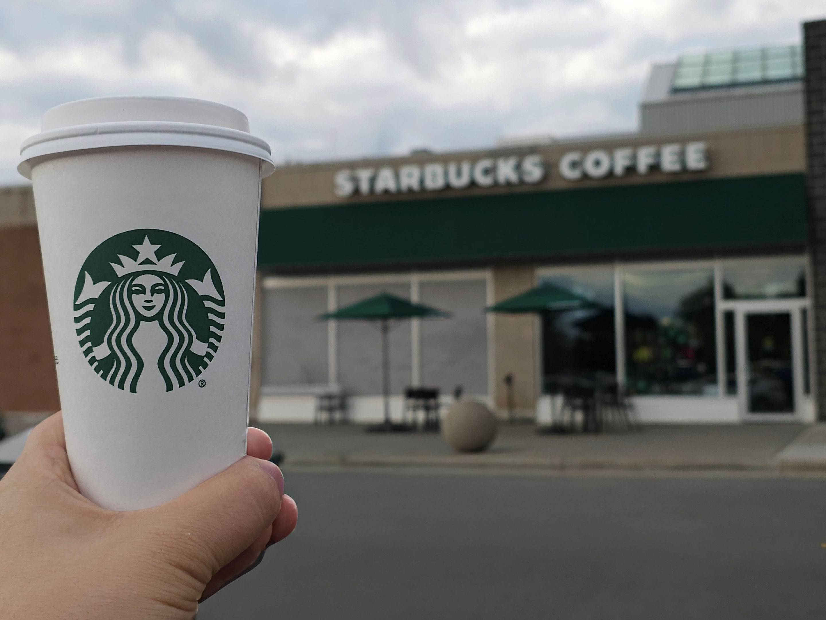 A hand holds a white Starbucks cup with the green logo in the foreground. In the background, a Starbucks Coffee storefront with a green awning, glass windows, and outdoor seating is visible under a cloudy sky.