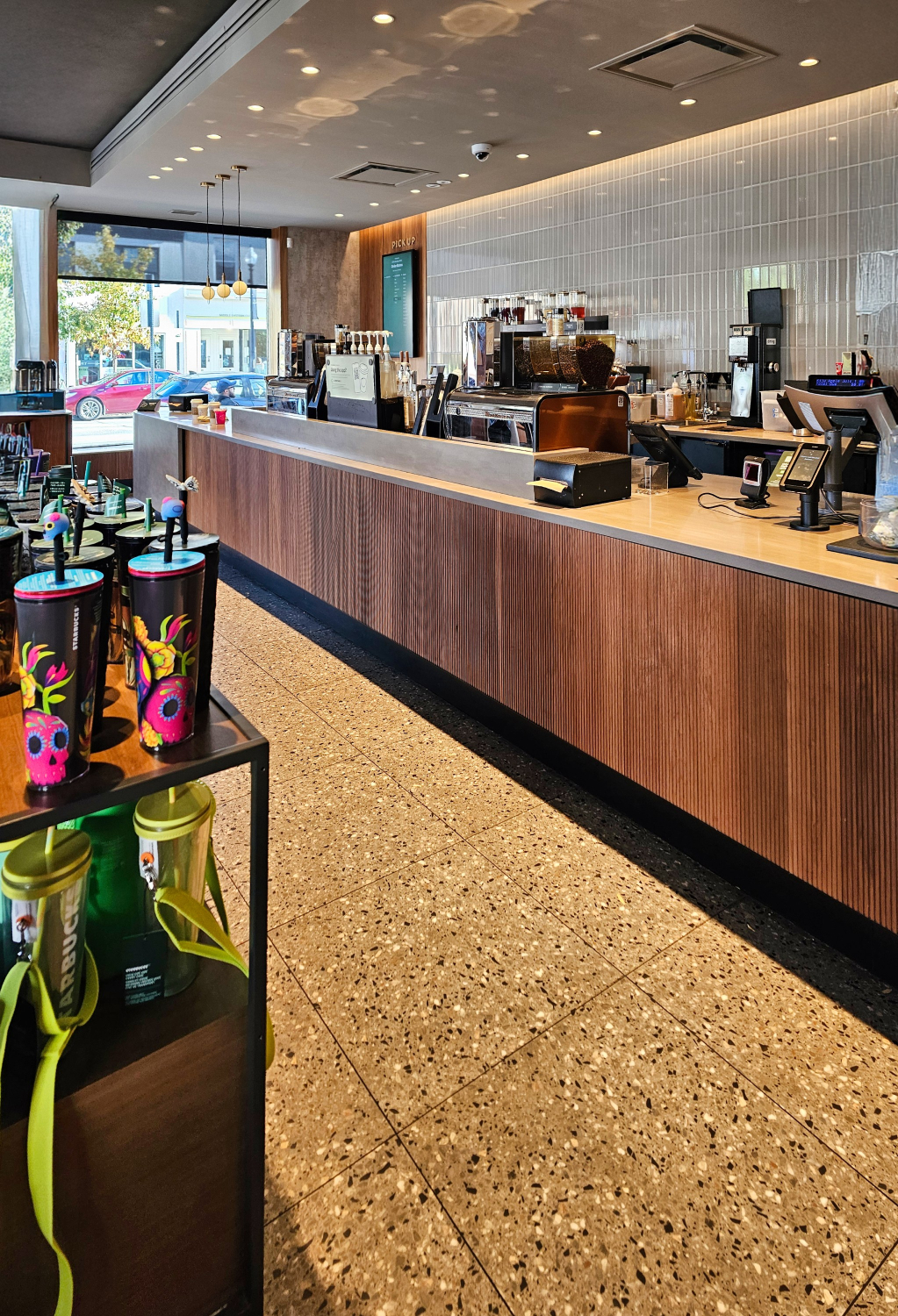 A modern café counter with espresso machines, payment terminals, and wood‑panelled finishes lines the space. Polished terrazzo flooring, tiled walls, and large front windows define the bright, contemporary interior.