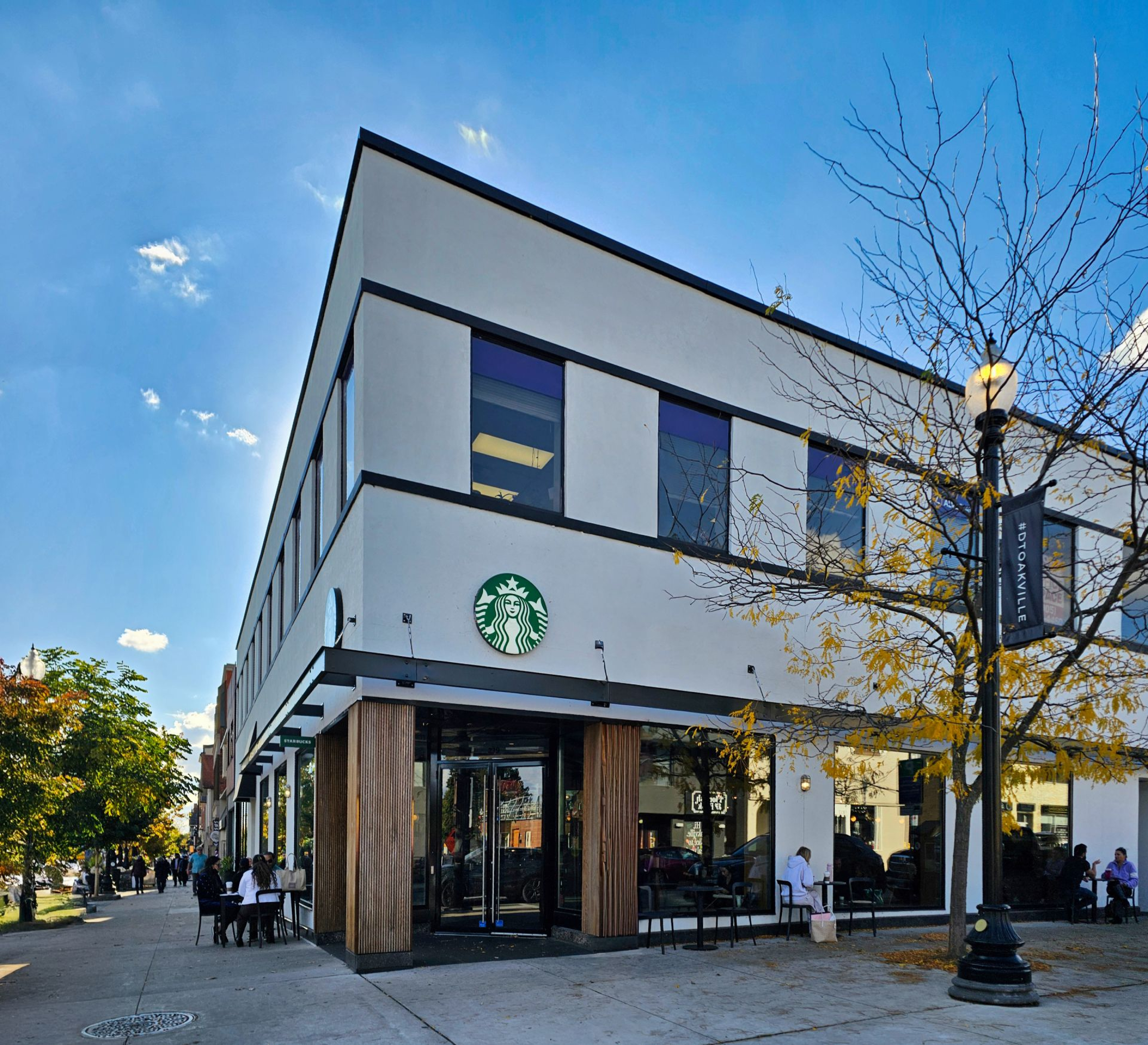 A corner Starbucks in a two‑storey building with a light-colored exterior, large windows, and wood column accents at the entrance. The green Starbucks logo is mounted above glass doors along a city sidewalk with trees and outdoor seating.