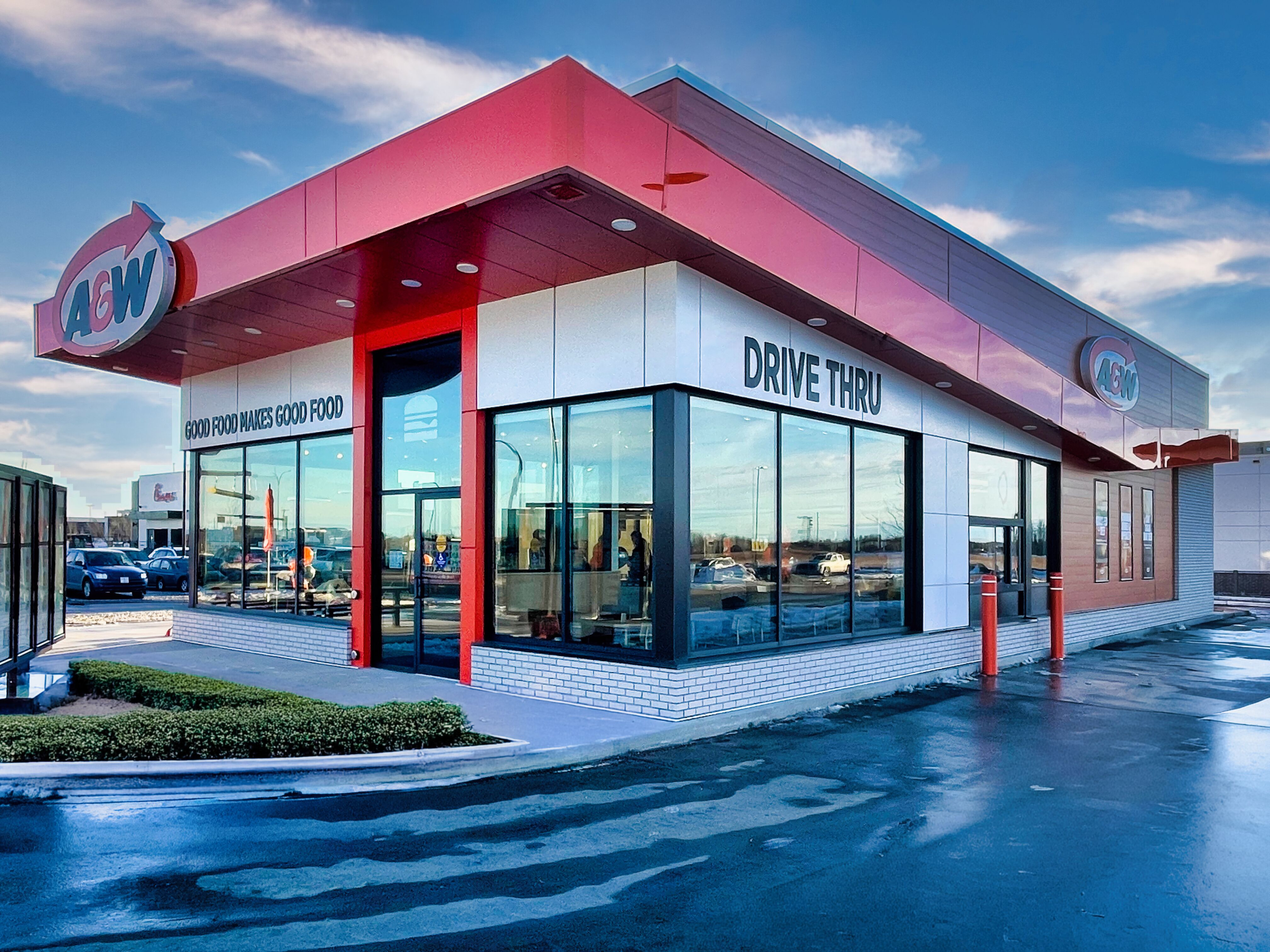 A modern A&W fast‑food restaurant with red and white exterior panels, large glass windows, and visible “Drive Thru” signage, shown on a clear day with a paved parking area in the foreground.