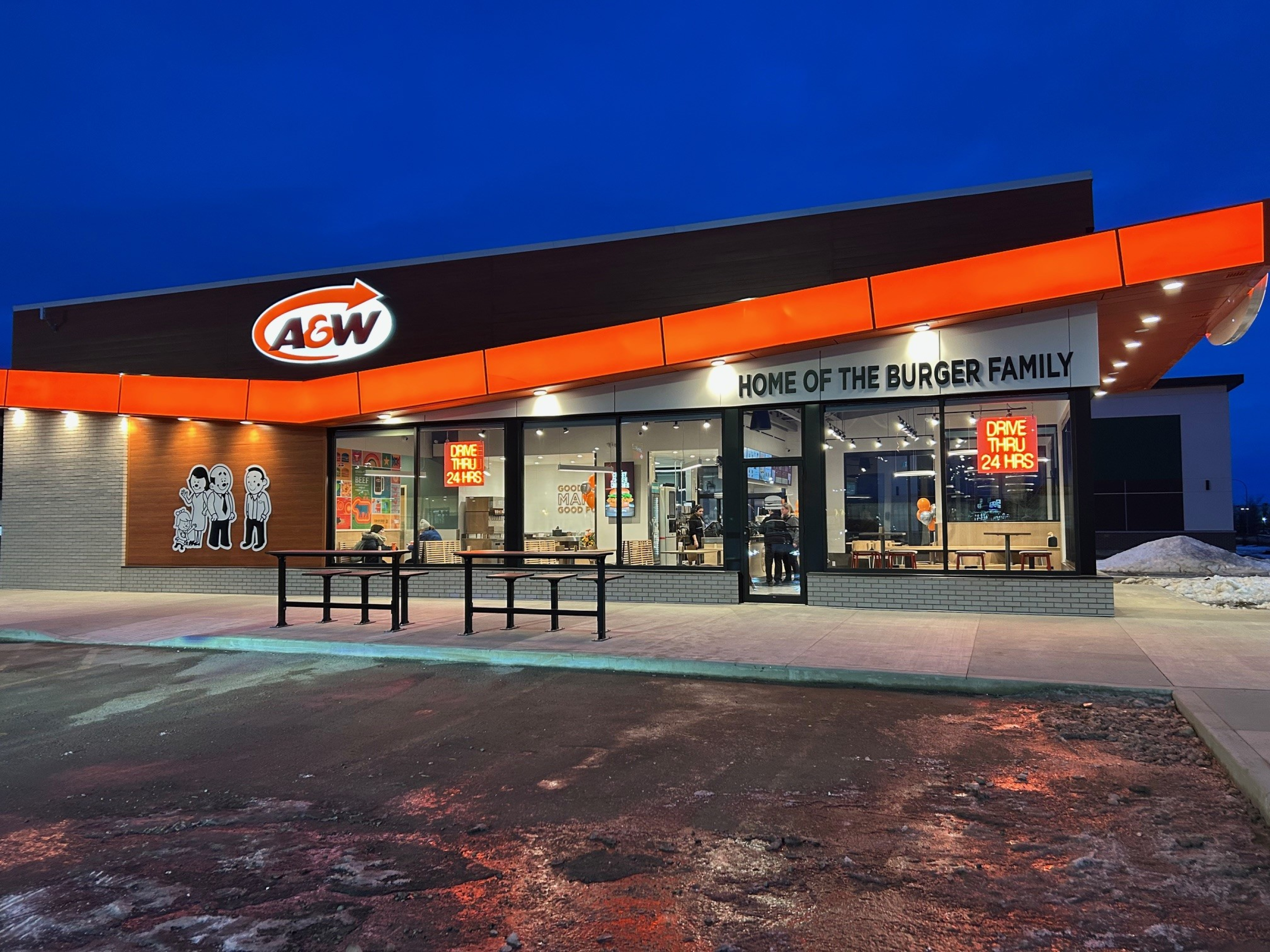 Exterior of an A\&W restaurant at dusk, featuring a modern orange-and-brown facade with illuminated A\&W signage, large glass windows, and outdoor seating in front of the building.