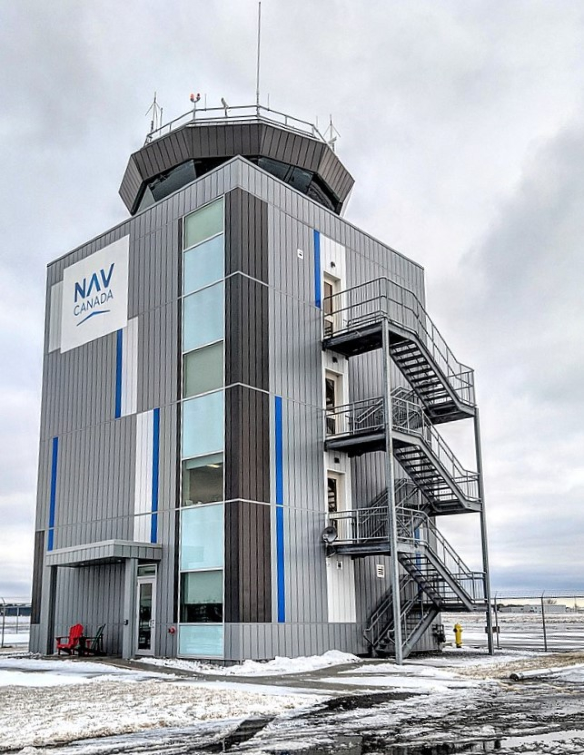 A NAV CANADA air traffic control tower with a gray metal exterior, vertical blue accents, and an exterior staircase is shown in a snowy setting. The tower stands within a fenced site under an overcast sky.