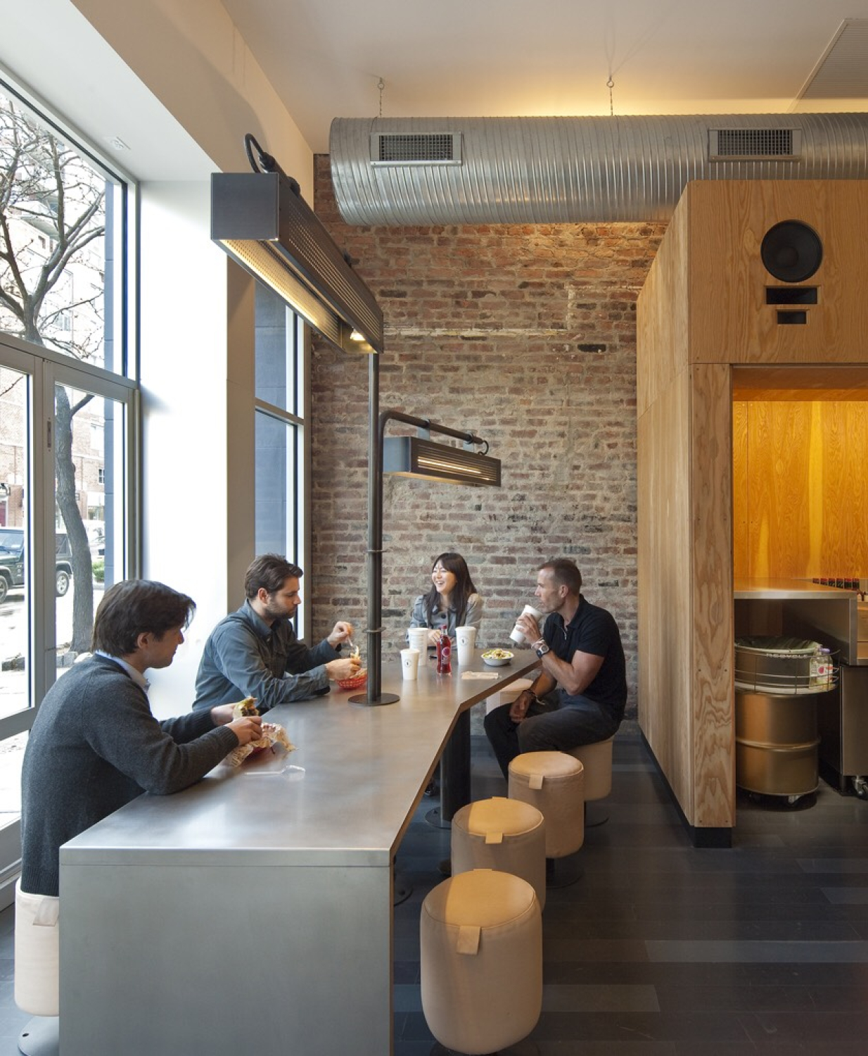 A casual dining area with people seated along a counter at a high table near large windows. Exposed brick, metal ductwork, wood accents, and pendant lights create a modern, industrial-style interior.