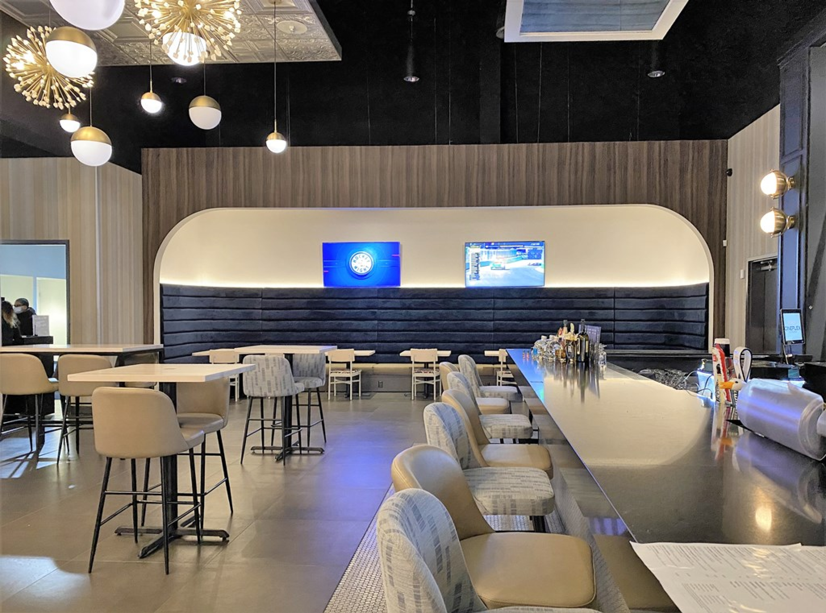 A Cineplex VIP lounge bar area featuring a long, polished bar with high‑backed stools and light wood tables nearby. Upholstered banquette seating, decorative pendant lights, and wall‑mounted screens create an upscale, modern cinema dining space.