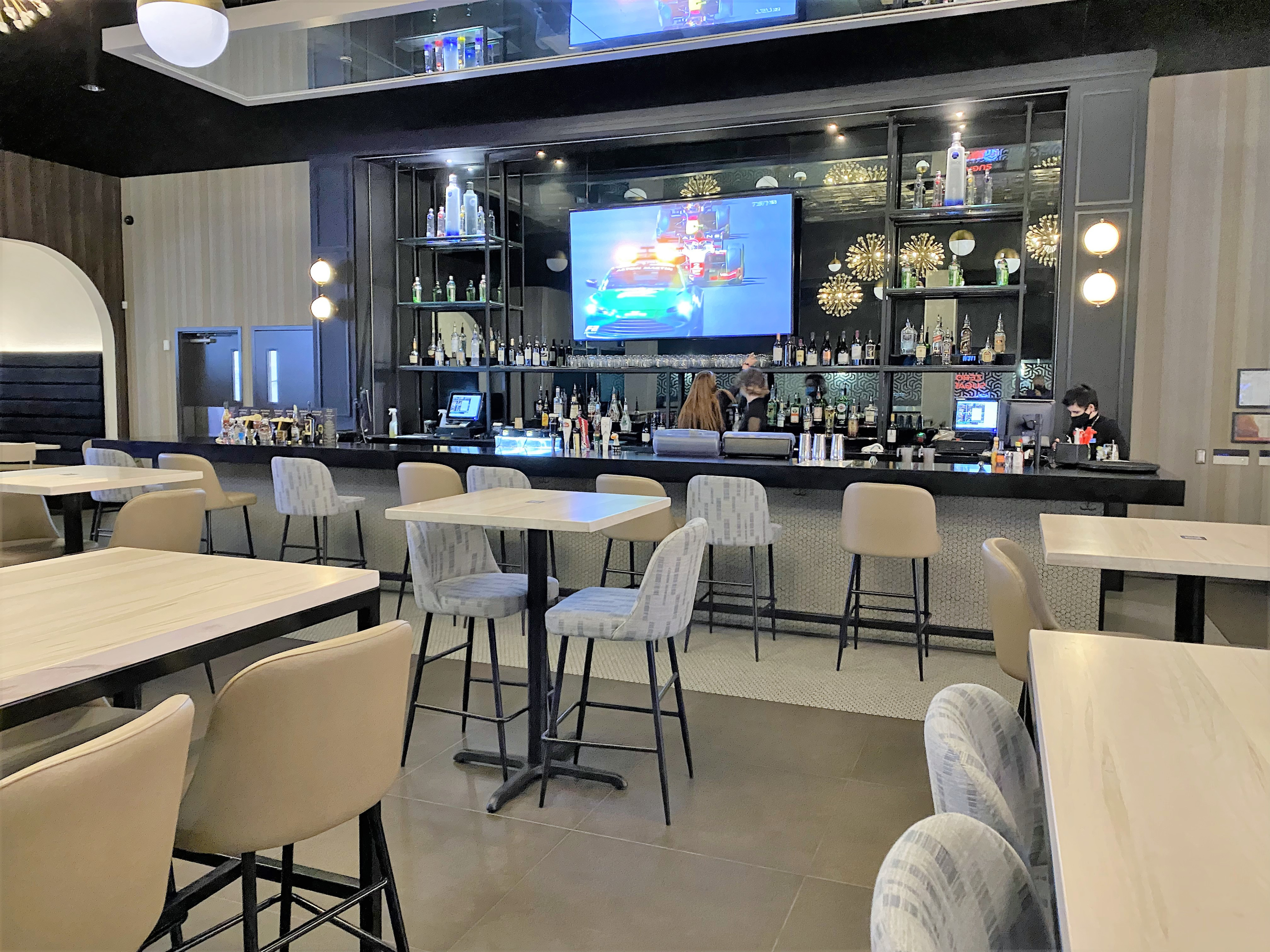A Cineplex VIP lounge bar with high‑top tables and upholstered stools arranged around a long bar counter. Shelving with bottles, decorative lighting, and a wall‑mounted screen create an upscale cinema dining atmosphere.