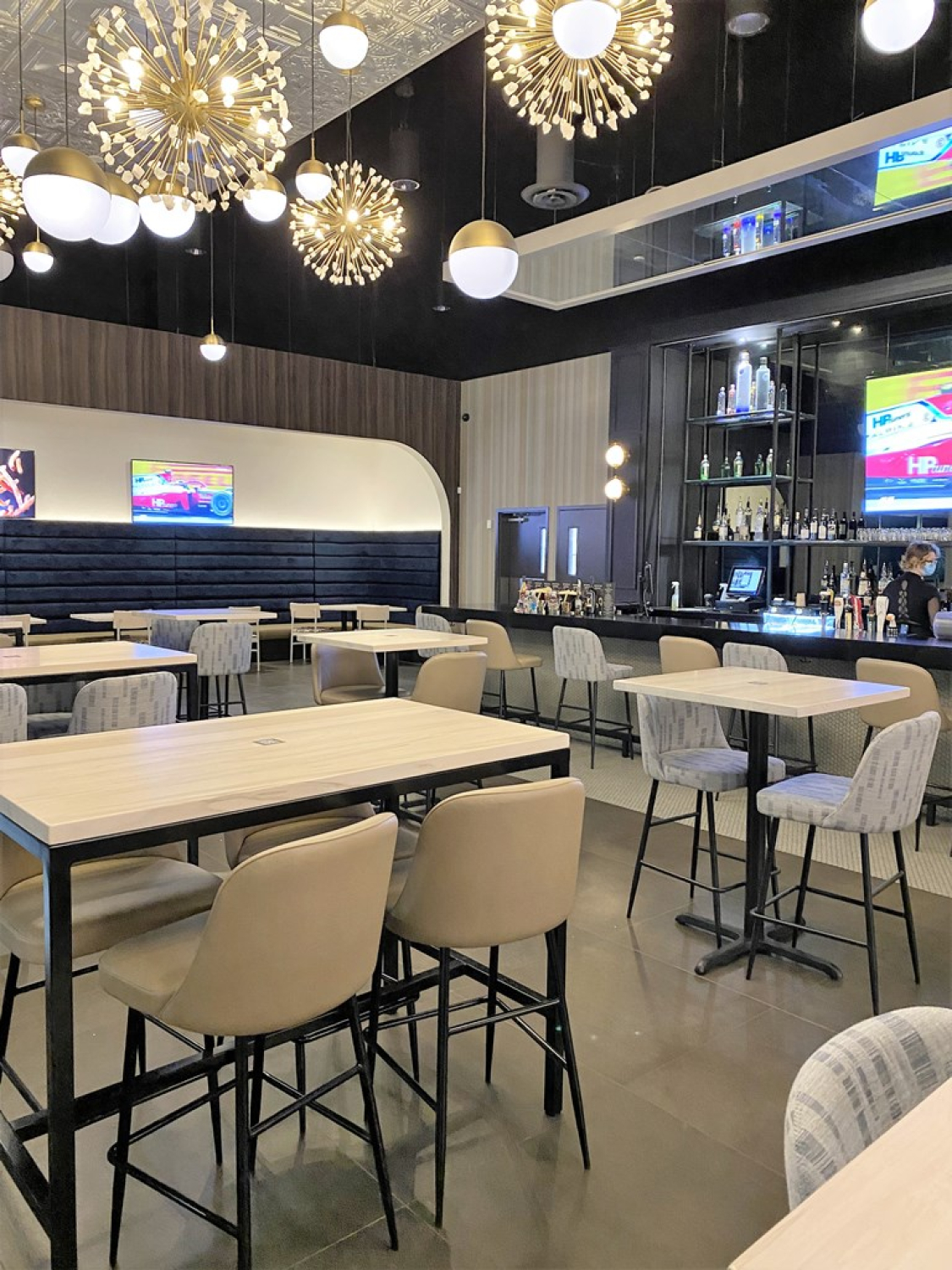 A Cineplex VIP lounge dining area with light wood tables, upholstered high‑top chairs, and a full bar along the back wall. Decorative pendant lights, mirrored ceiling panels, and wall‑mounted screens create a modern, upscale cinema atmosphere.