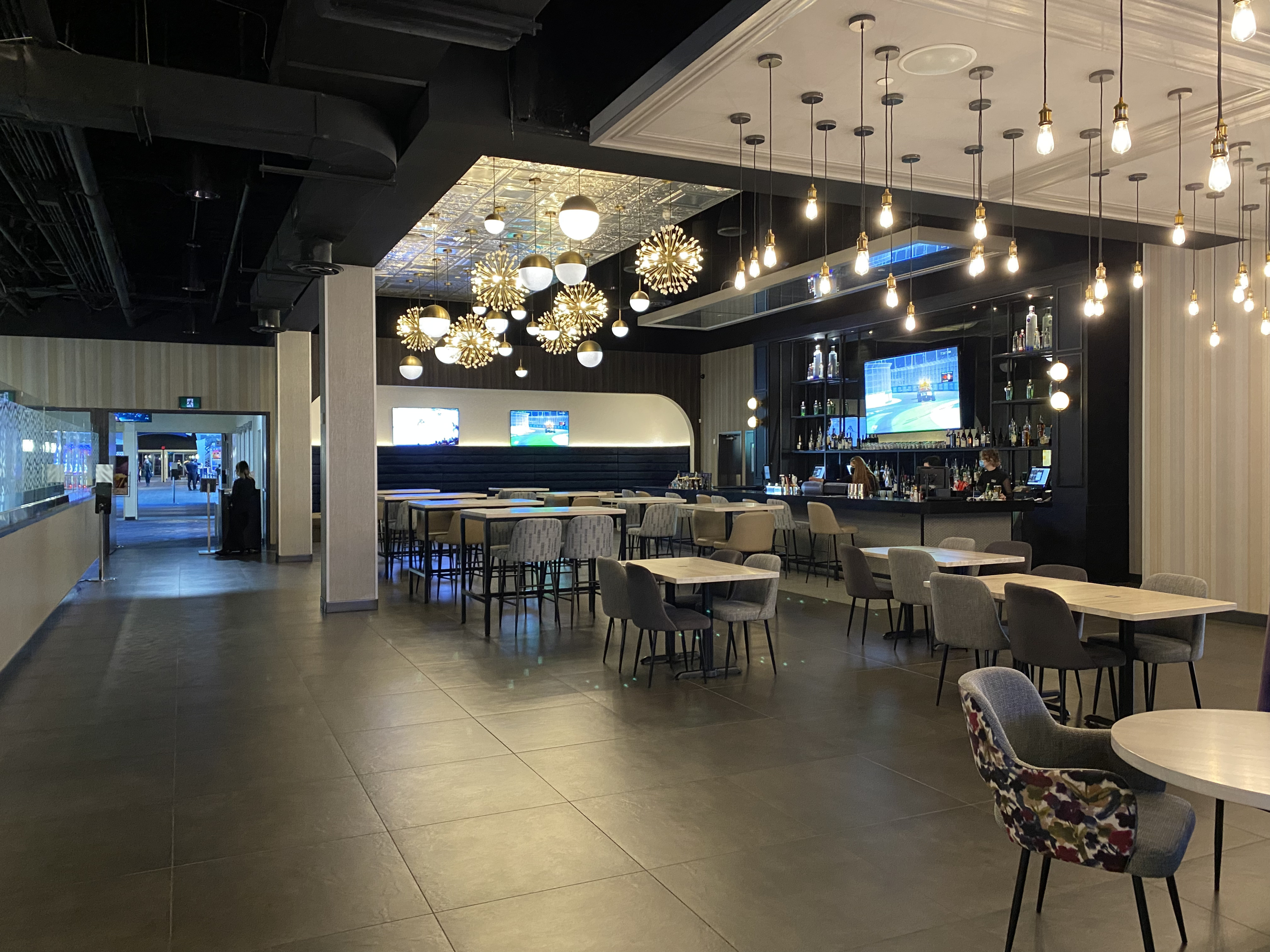 A Cineplex VIP lounge dining area with modern tables, upholstered chairs, and a long bar along the back wall. Decorative pendant lights, reflective ceiling panels, and wall‑mounted screens create an upscale, open pre‑theatre atmosphere.