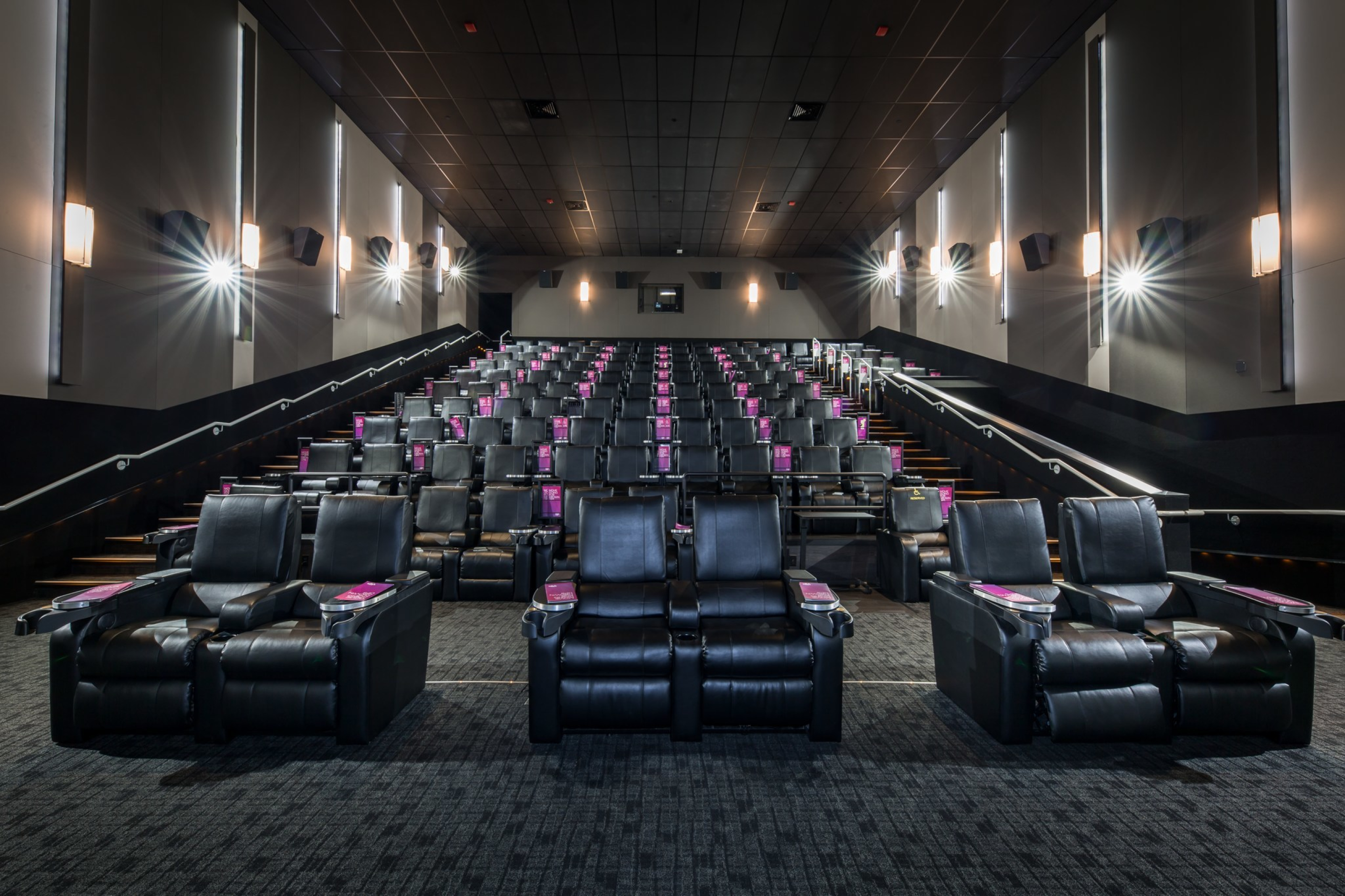 A Cineplex VIP cinema auditorium with rows of wide black reclining seats arranged in tiered seating facing a large screen. Soft wall lighting, carpeted floors, and wide aisles create an upscale theatre environment.