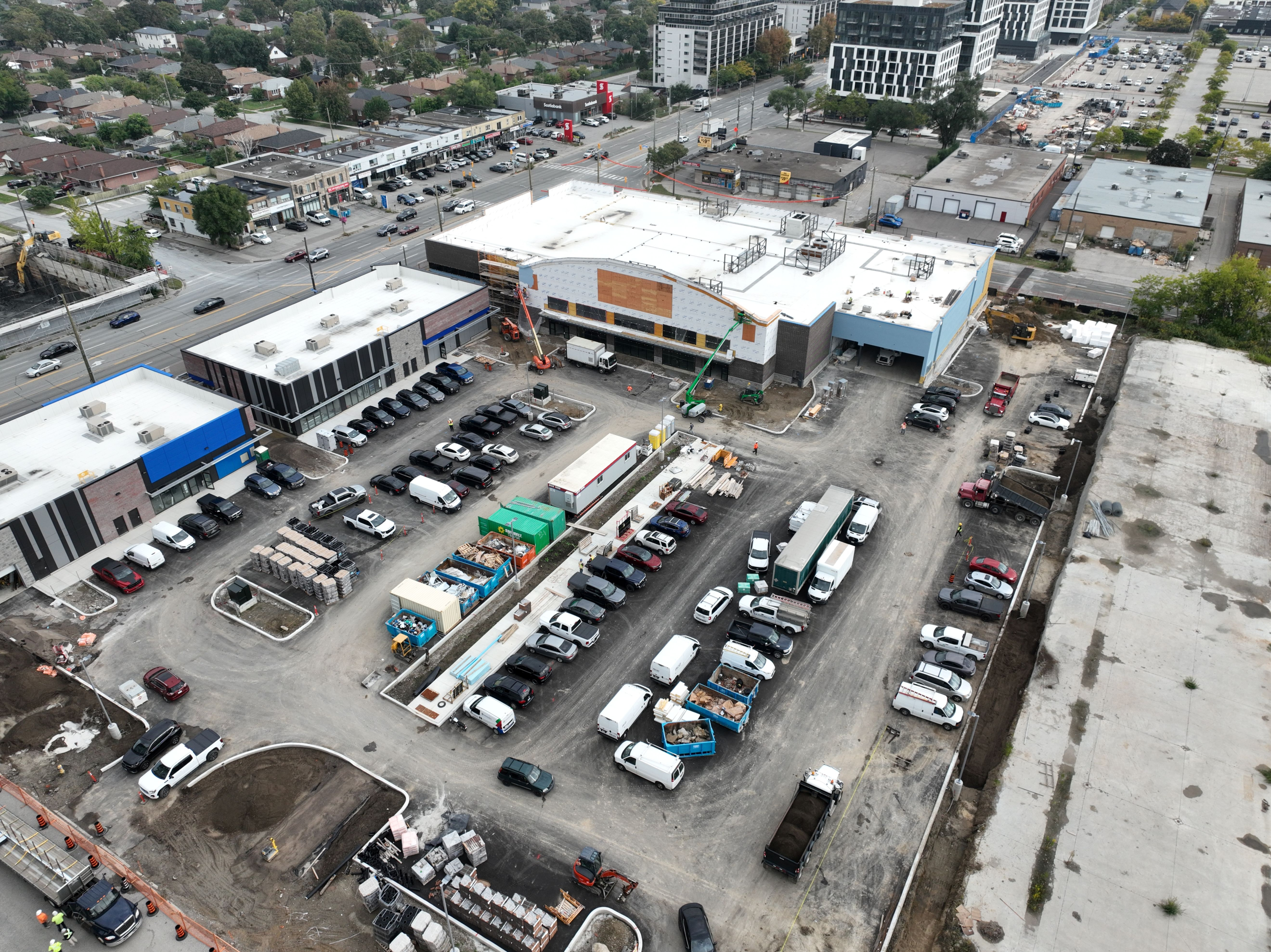 An aerial view of a commercial plaza under construction, showing multiple low‑rise buildings with white roofs, surrounding parking areas, construction vehicles, and materials, with nearby roads, shops, and houses visible around the site.