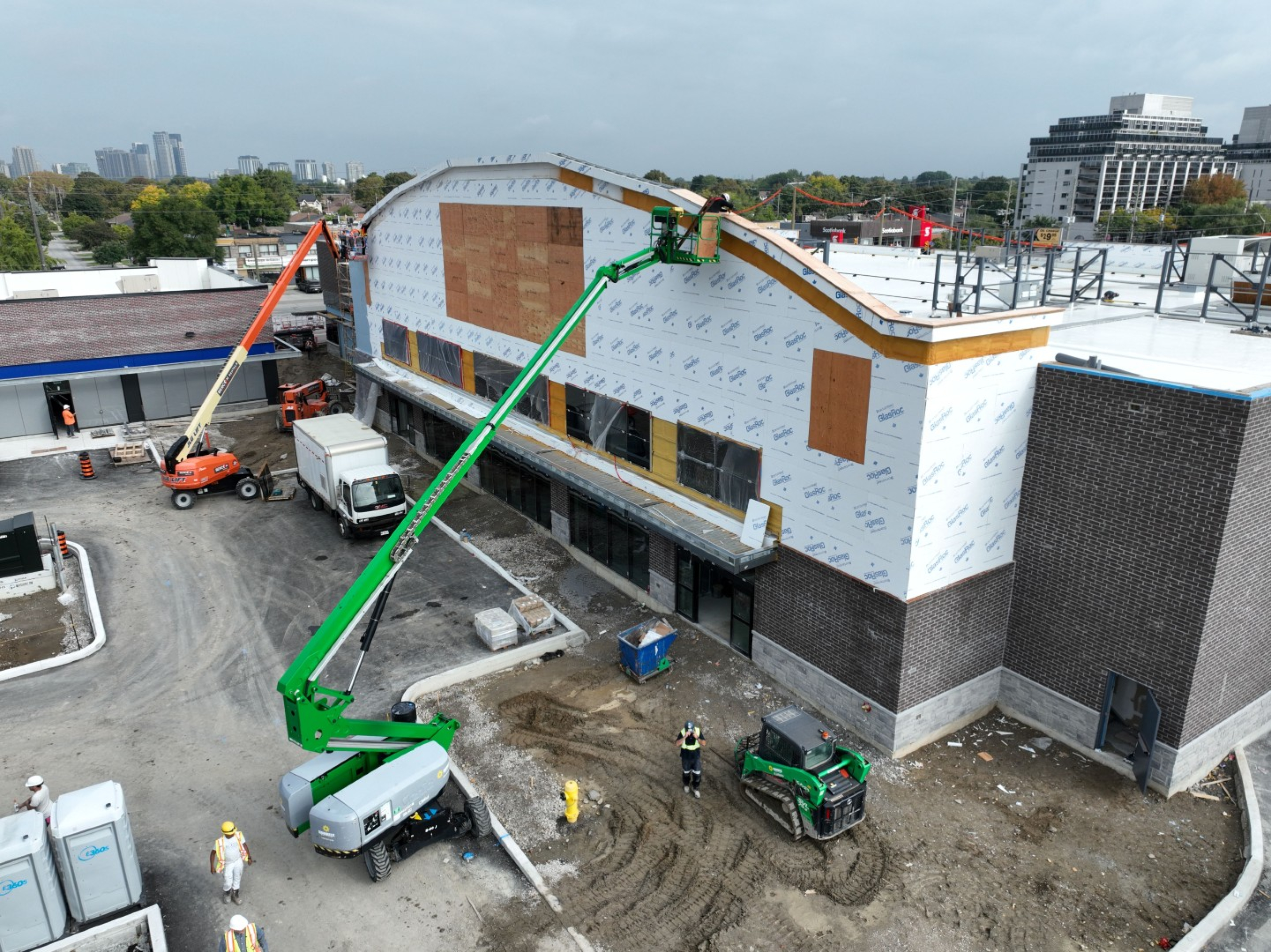 An angled aerial view of a commercial building under construction shows exterior walls covered in wrap, two boom lifts raised to the roofline, workers on site, and construction vehicles and materials on the ground below.