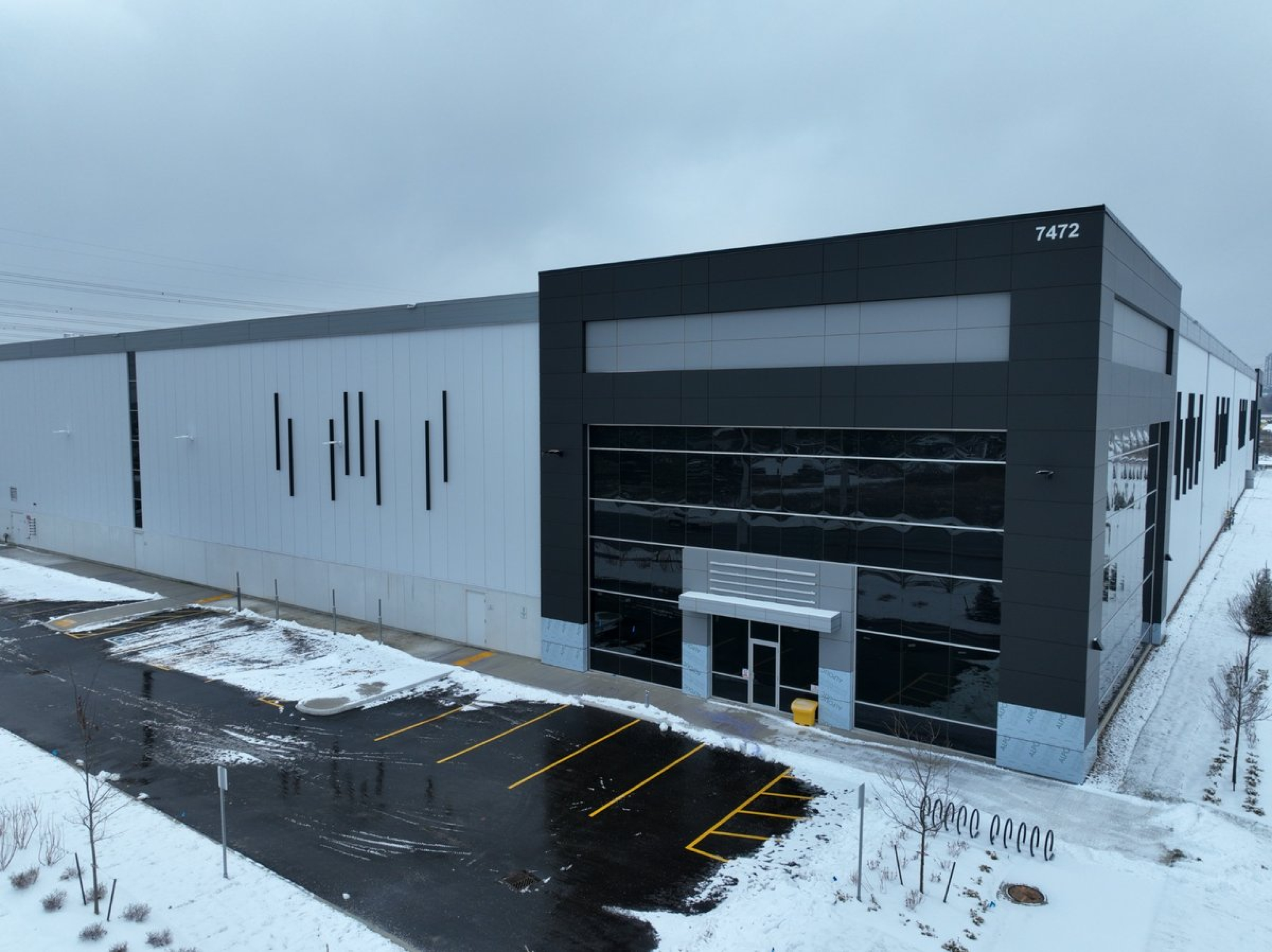 A modern industrial building with a white and dark gray exterior, shown from an elevated angle during winter. Snow covers the surrounding ground and parking areas, with a glass-fronted entrance visible on the facade.