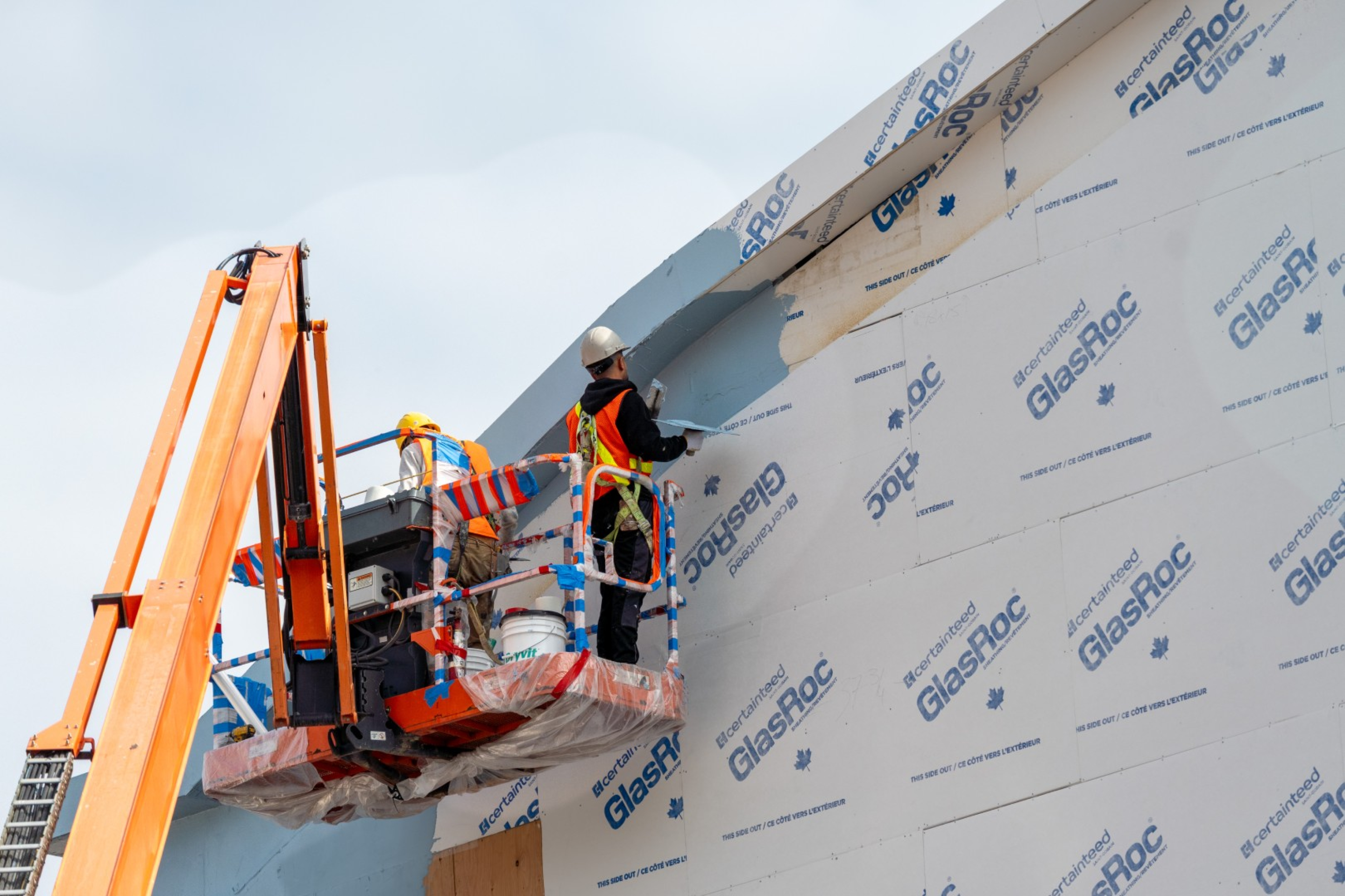 Two construction workers on a raised lift apply exterior cladding to a commercial building wall. Insulation panels, safety equipment, tools, and a bucket are visible against a clear sky.