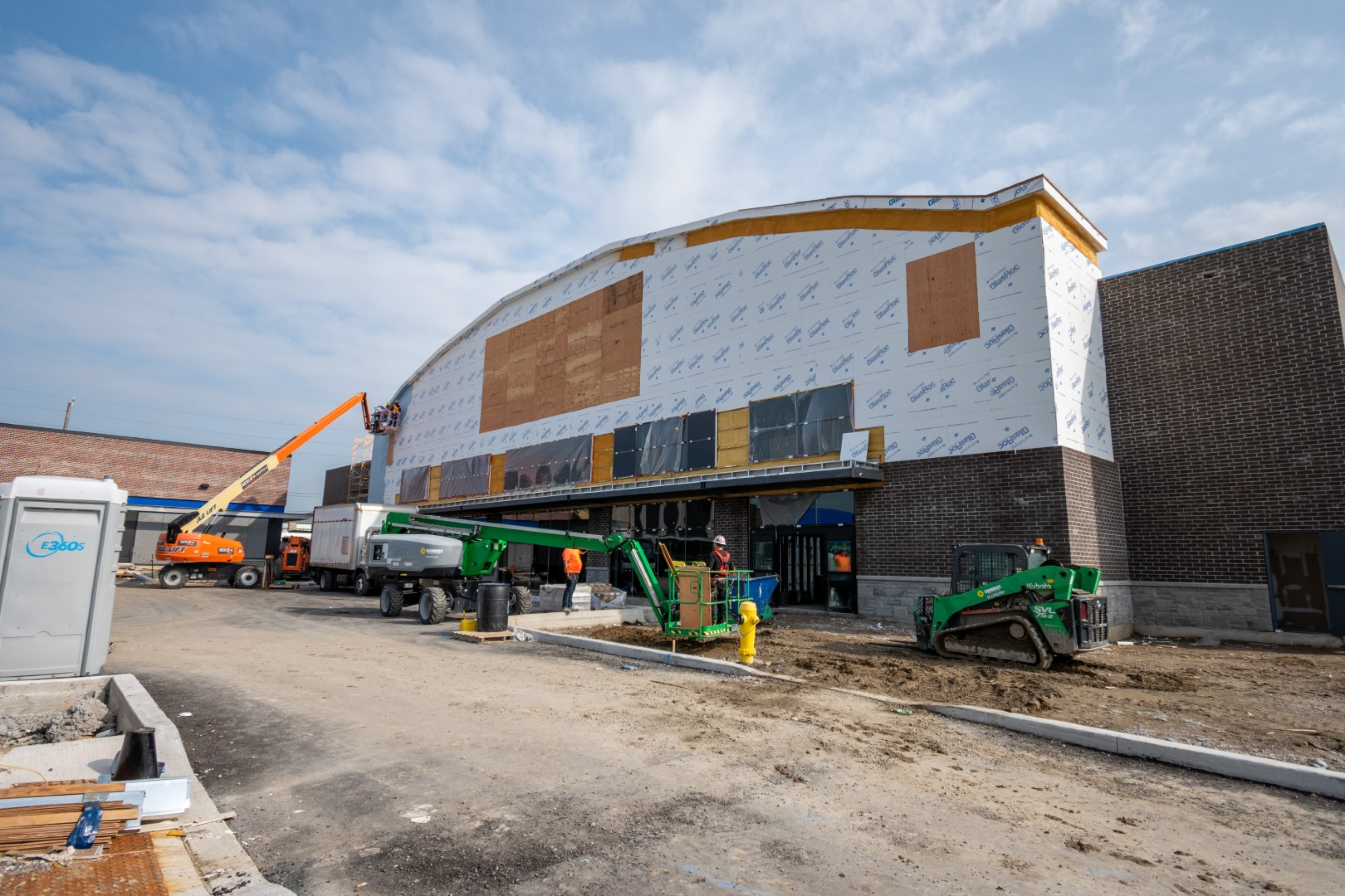 A commercial building under construction with exterior walls partially covered in insulation wrap. Boom lifts, construction equipment, workers, and muddy ground are visible along the unfinished facade under a bright sky.