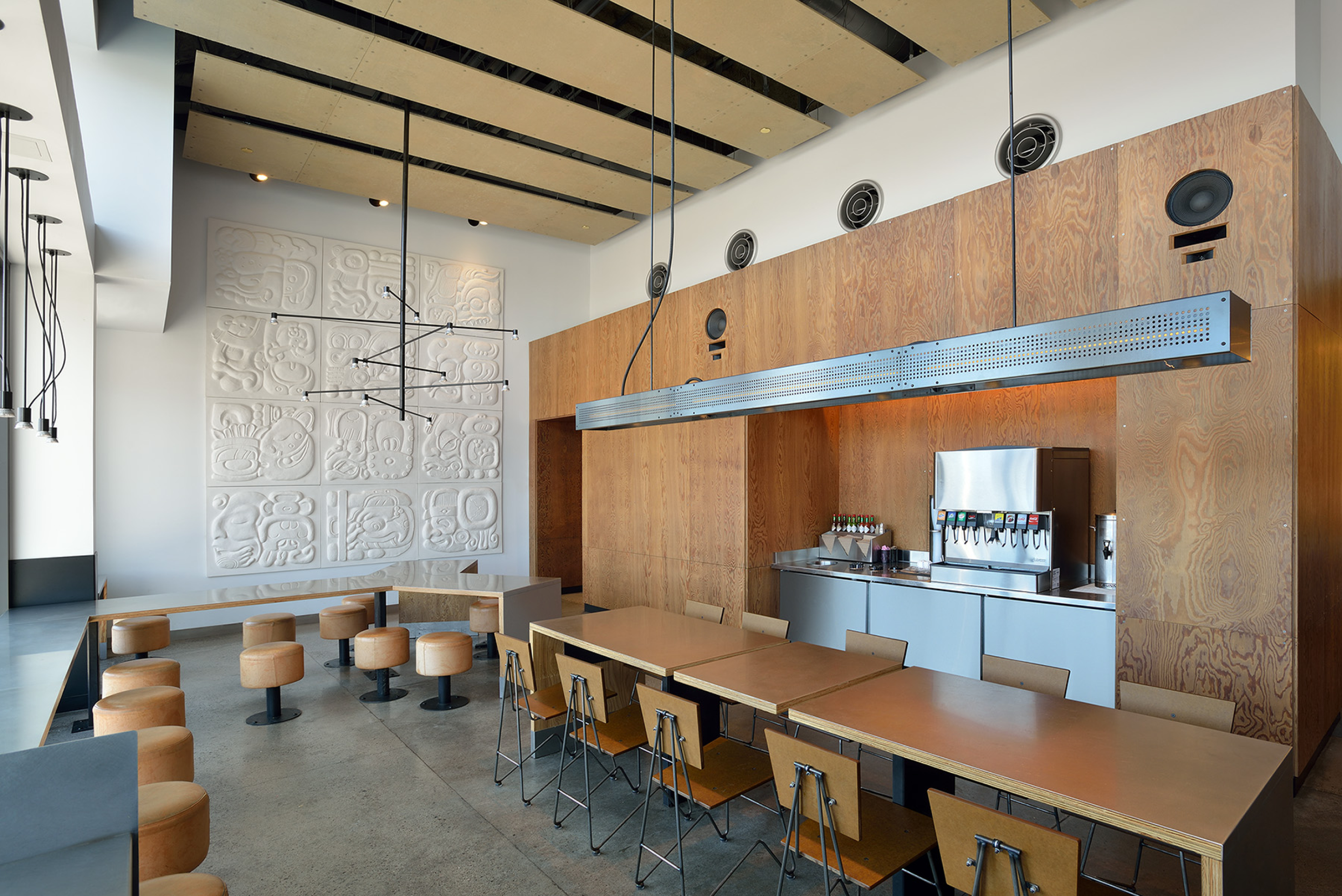 A modern restaurant seating area with long communal tables, wooden stools, and light wood wall panels featuring built-in beverage dispensers. Decorative wall art, hanging lights, and exposed ceiling elements create a clean, contemporary interior.