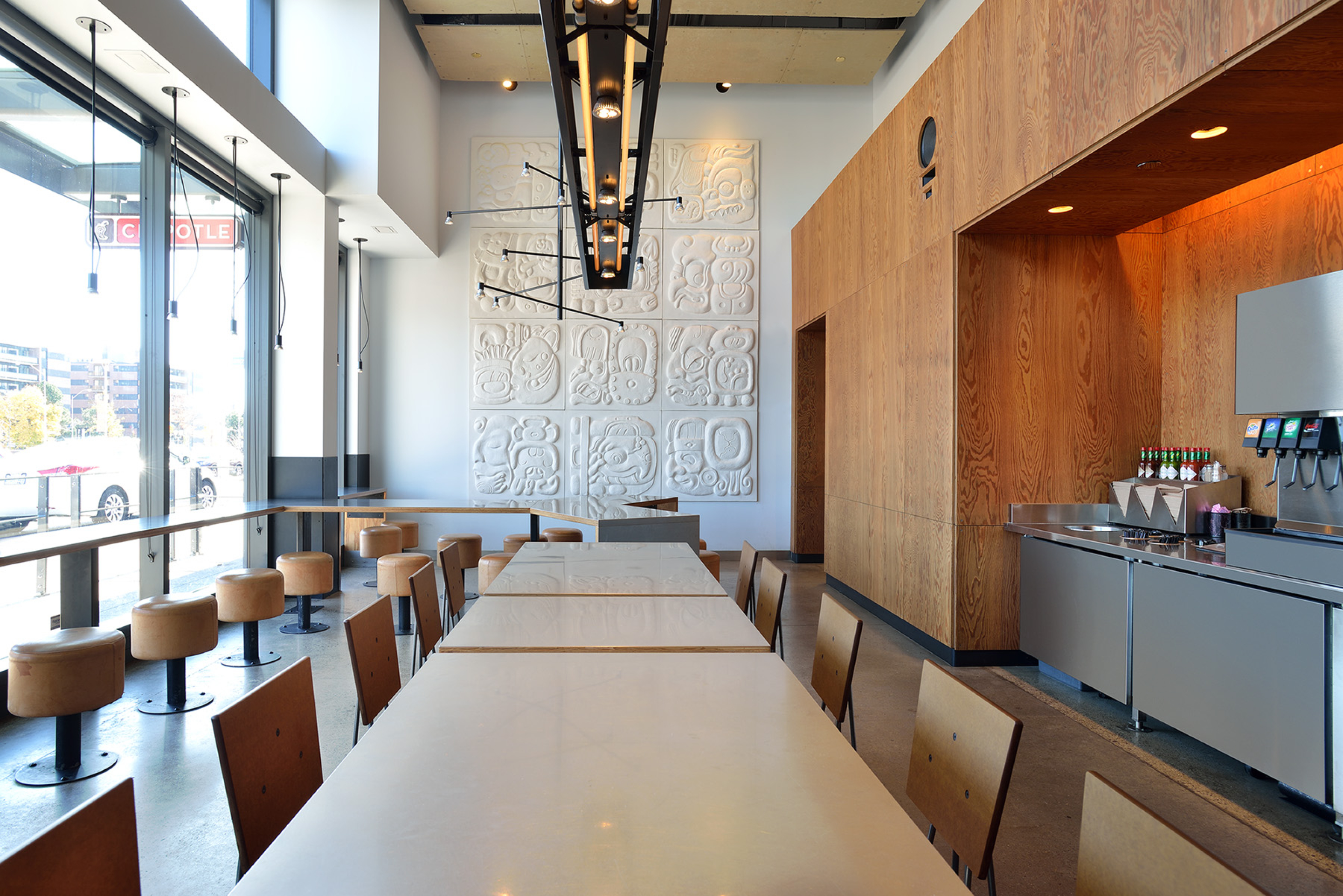 A bright Chipotle dining area with long communal tables, wooden chairs, and round stools beside large windows. Light wood wall panels, a beverage station, decorative wall art, and hanging lights create a modern interior.