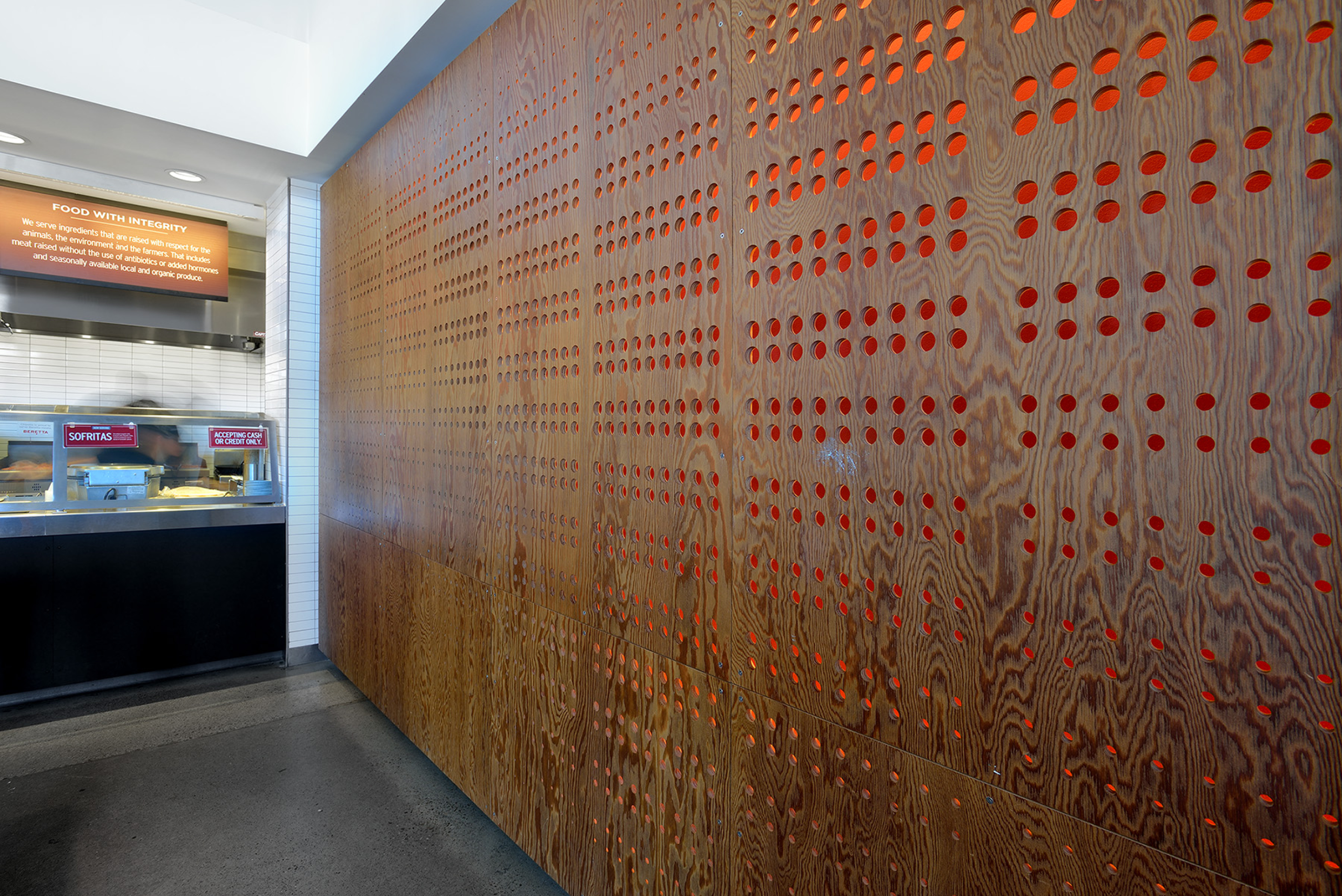 A Chipotle interior featuring a wood-paneled wall with a grid of small circular cutouts backed by orange accents. Part of the counter area and menu signage is visible to the left under bright lighting.