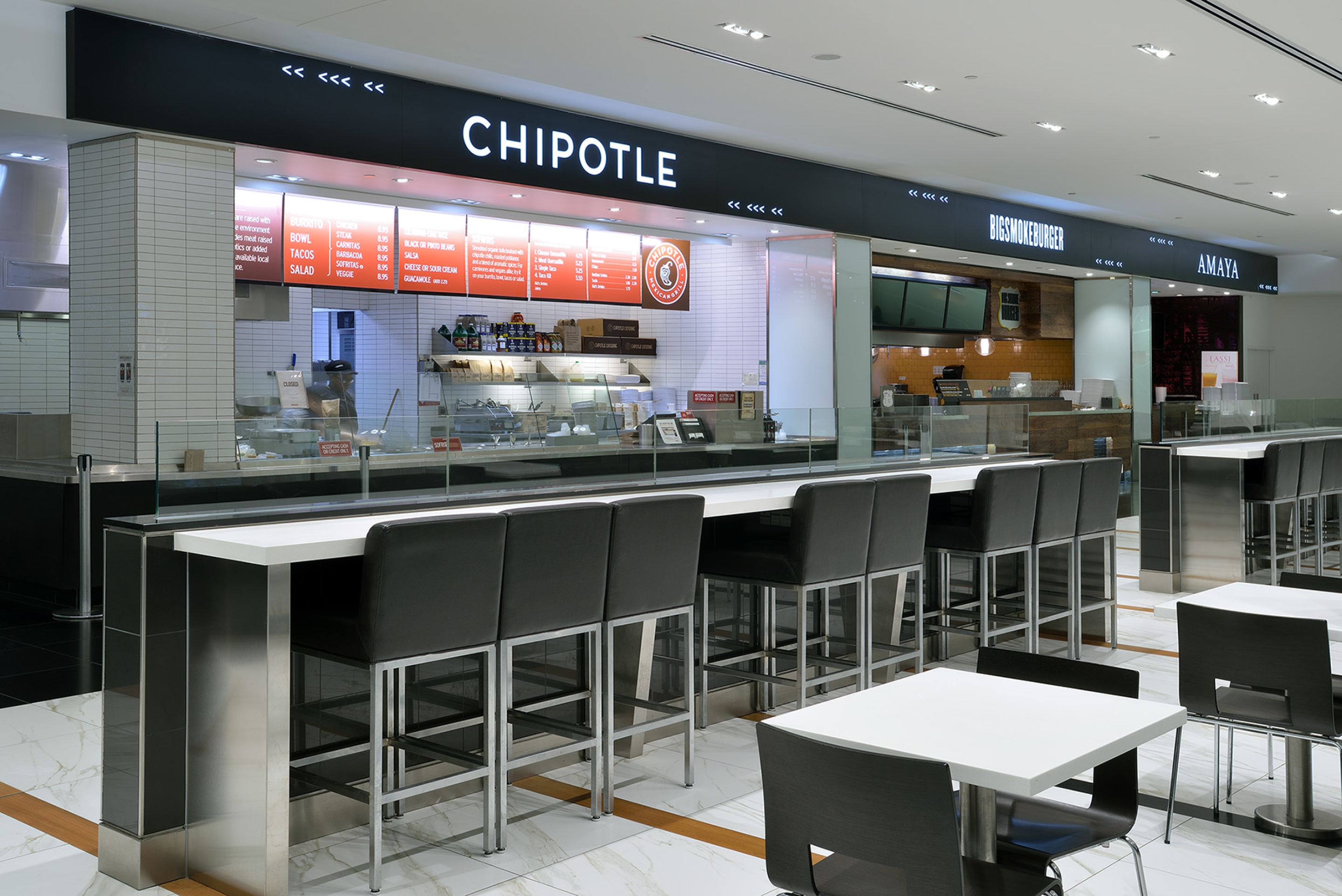 A Chipotle restaurant counter inside a food court, with menu boards, glass display cases, and a row of high stools at a bar-height counter. Stainless steel finishes, bright lighting, and tiled walls create a clean, modern setting.