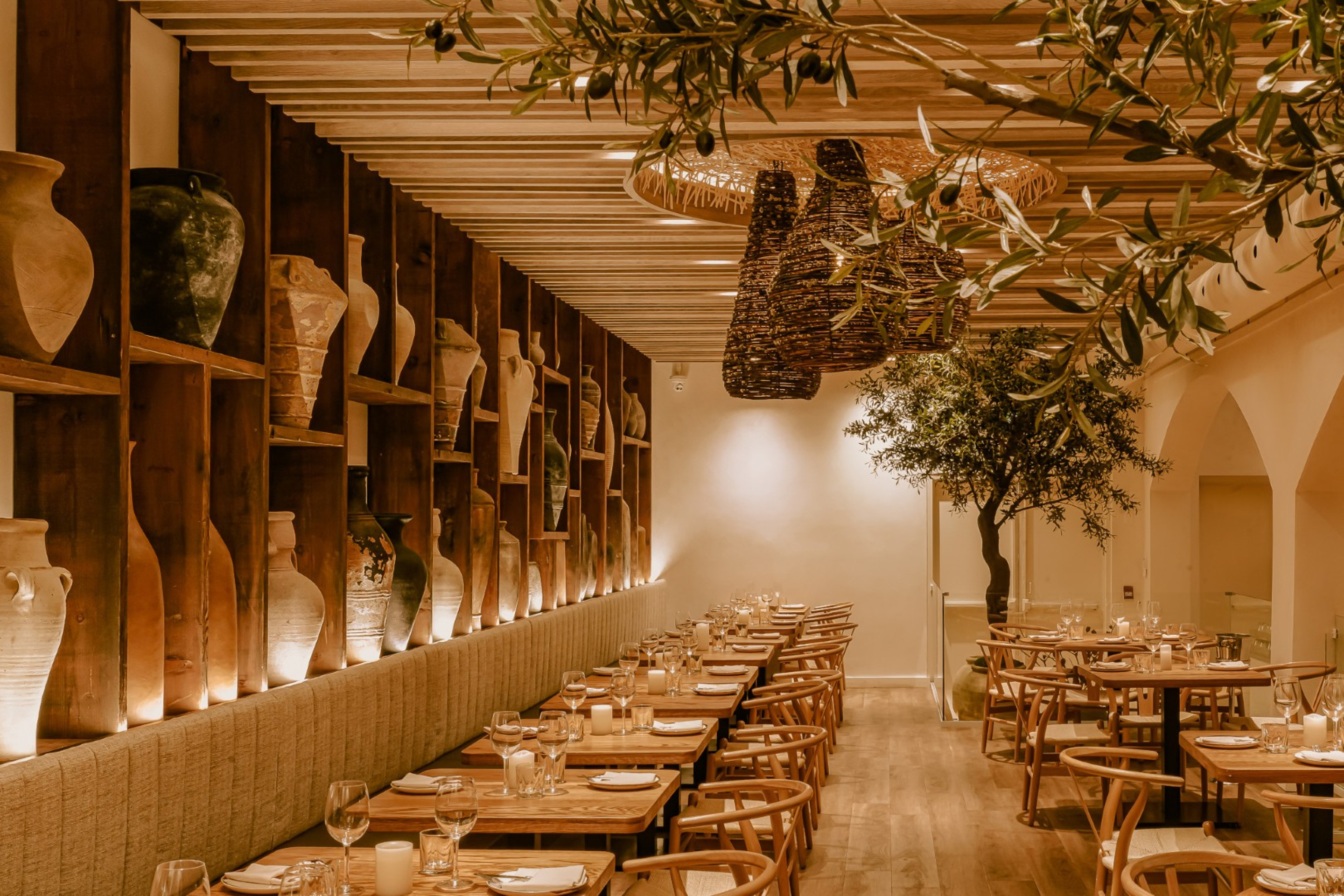 A warm, modern restaurant dining area with wood tables and chairs arranged beneath a slatted wood ceiling. Decorative pottery shelves, hanging woven lights, and indoor greenery create a natural, inviting atmosphere.