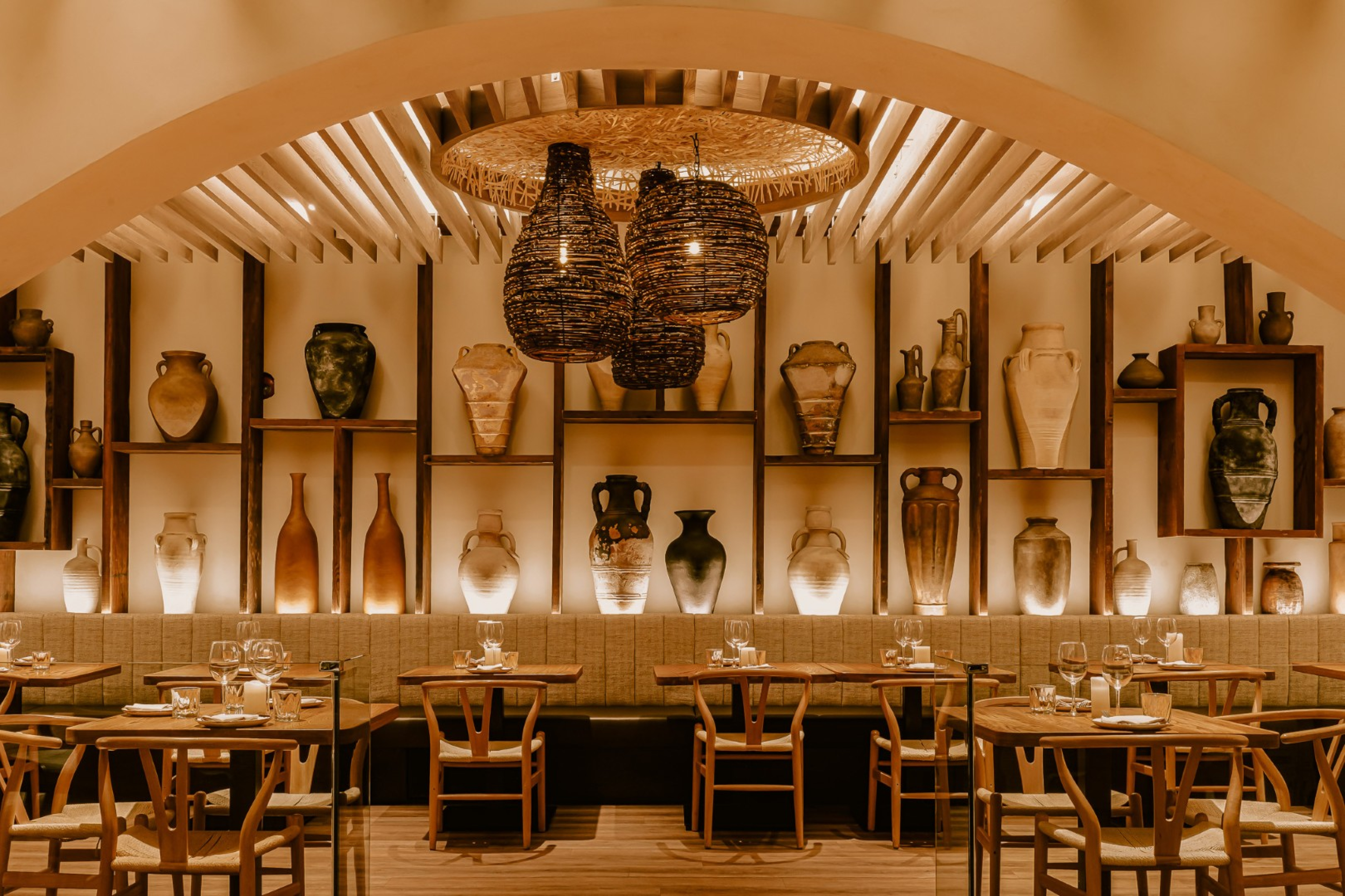 A warmly lit restaurant dining area with wood tables and chairs arranged beneath an arched, slatted wood ceiling. Shelves of decorative pottery line the wall, with woven pendant lights and place‑set tables creating an elegant atmosphere.