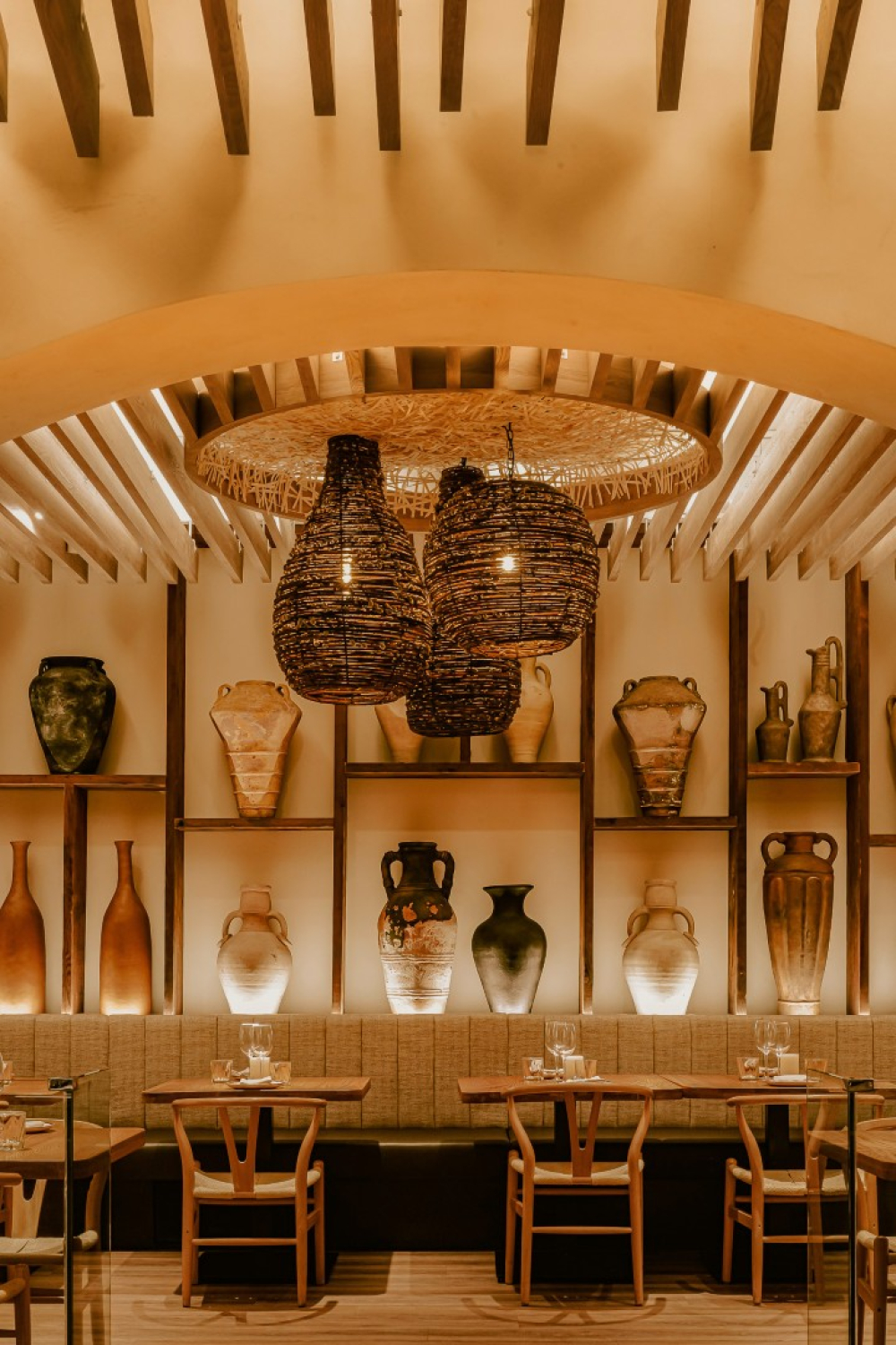 A warmly lit restaurant interior with wood tables and chairs arranged beneath an arched, slatted ceiling. Decorative pottery displayed on wall shelves and woven pendant lights create a cozy, upscale dining atmosphere.