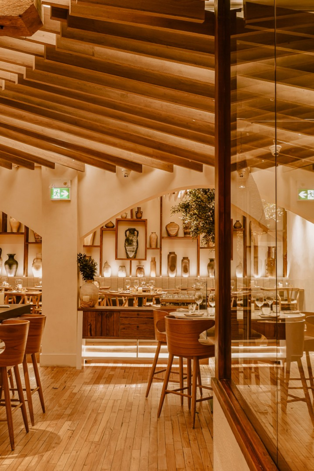 A warm, wood‑accented restaurant interior with tables and chairs arranged beneath exposed ceiling beams. Decorative pottery shelves, indoor plants, and soft lighting create an inviting, modern dining space.