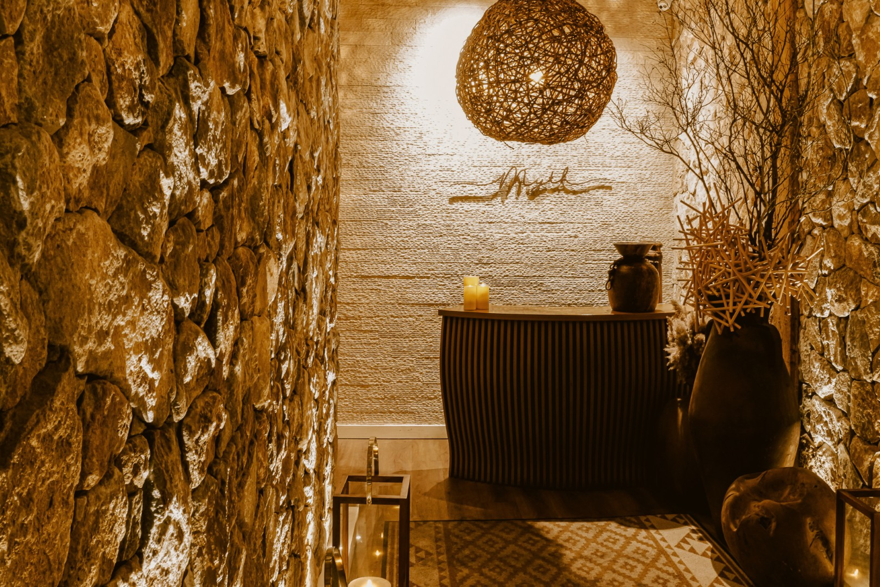 A softly lit entry alcove with textured stone walls leading to a small host stand. Woven pendant lighting, decorative vases, candles, and sculptural branches create a warm, intimate restaurant atmosphere.