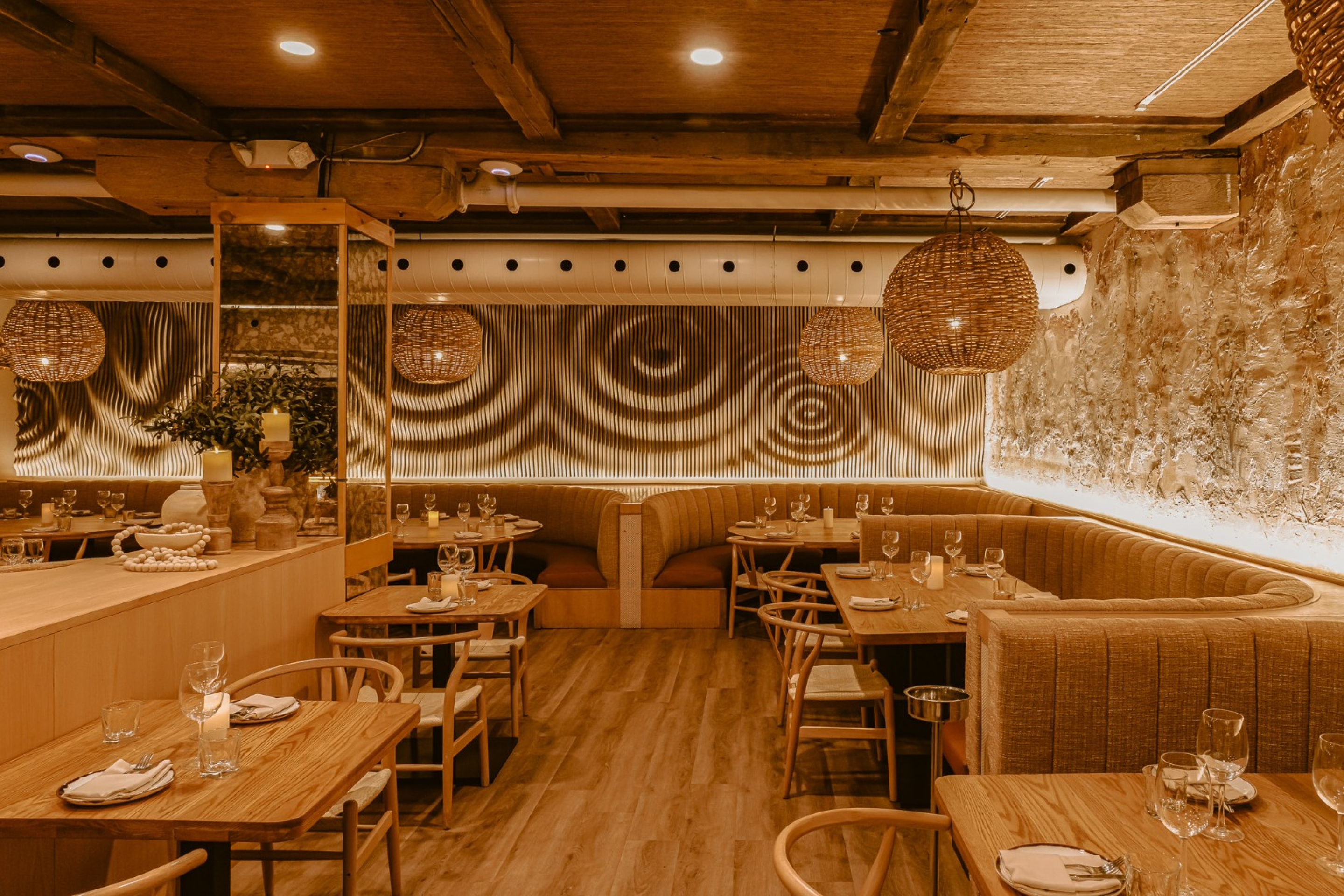 A warm, modern restaurant dining area with wood tables, tan upholstered booths, and woven pendant lights. Exposed ceiling beams, textured walls, and softly patterned lighting create an inviting, upscale atmosphere.