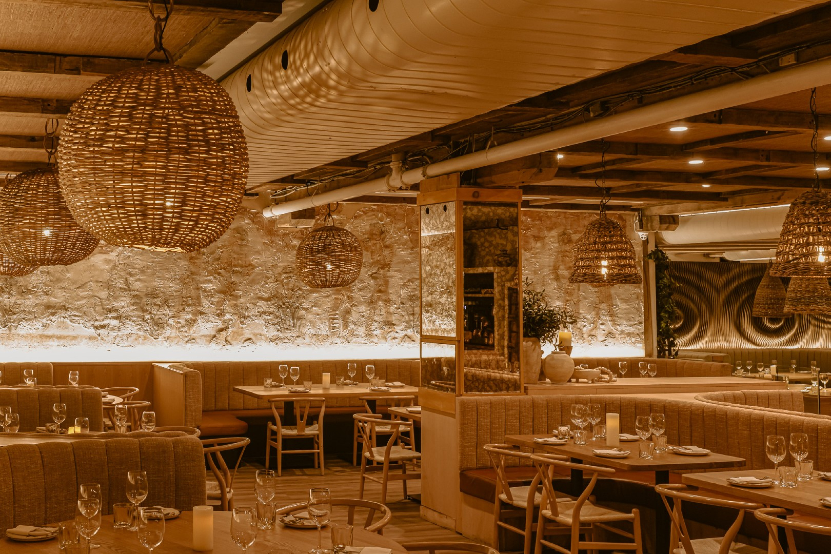 A warmly lit restaurant dining area with wood tables, cushioned booth seating, and woven pendant lights. Stone walls, exposed wood beams, and soft accent lighting create a cozy, upscale atmosphere.