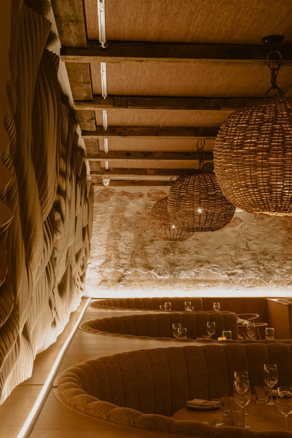 A warm restaurant interior with curved booth seating beneath woven pendant lights and exposed wood ceiling beams. Textured stone and sculpted wall panels line the space, softly lit to create an intimate dining atmosphere.