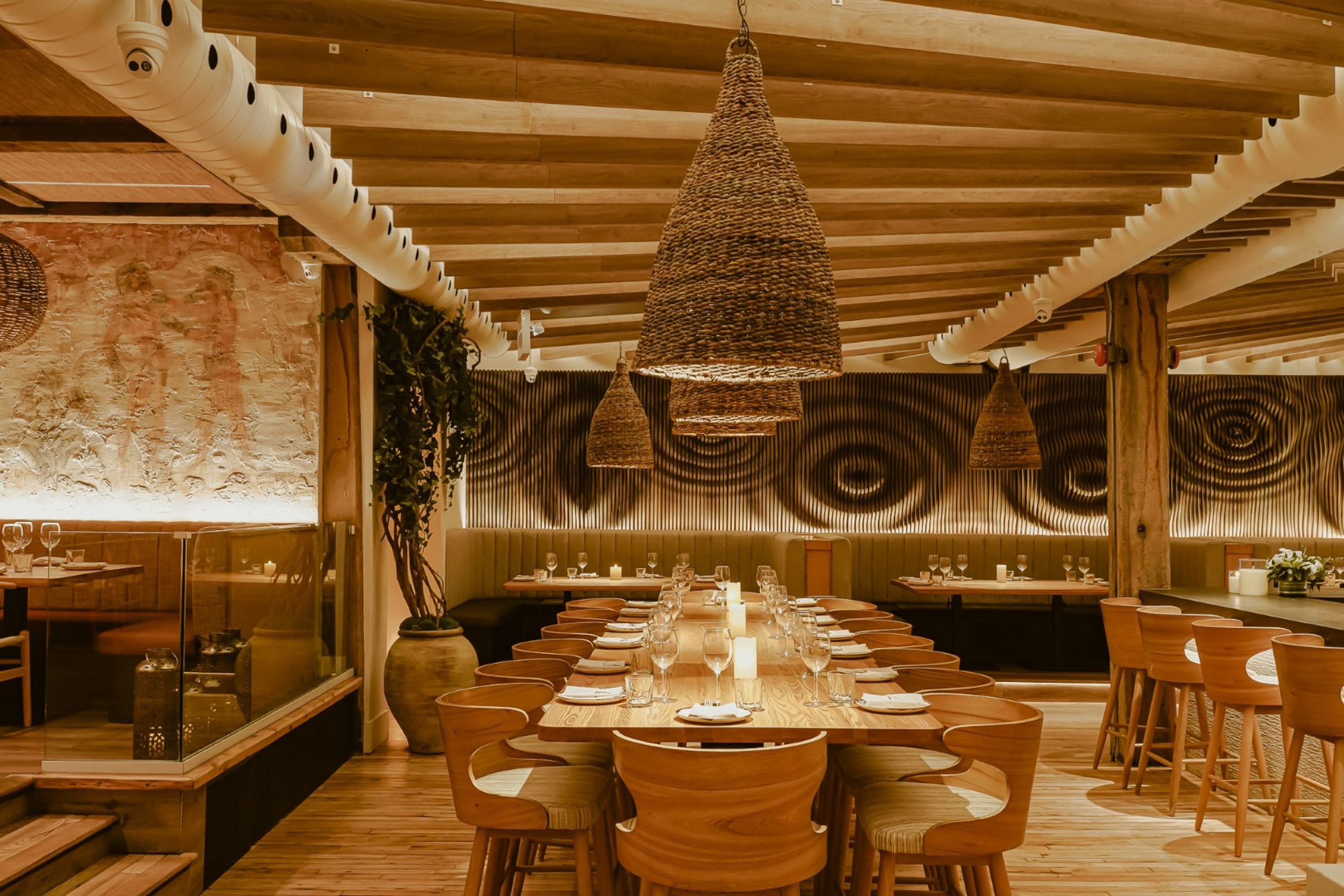 A warmly lit restaurant dining area with a long communal wood table set beneath exposed ceiling beams and woven pendant lights. Patterned wall panels, booth seating, and place settings create a refined, inviting atmosphere.