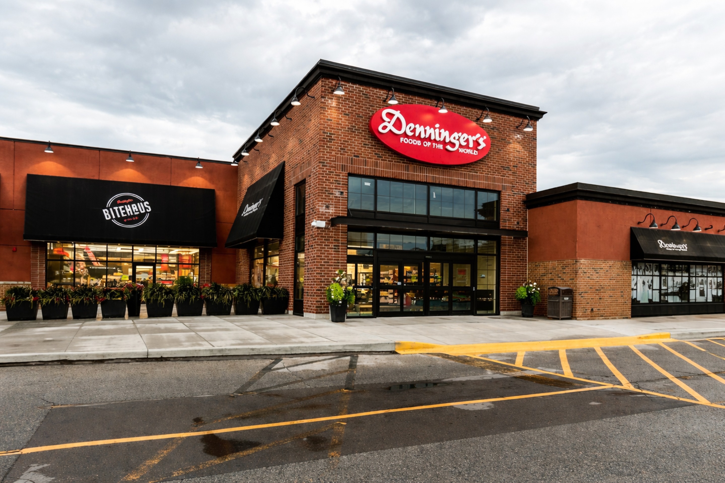 A Denninger’s grocery store exterior with a brick facade and a red “Denninger’s Foods of the World” sign above the main entrance. Large glass doors, storefront windows, and planters line the sidewalk under an overcast sky.