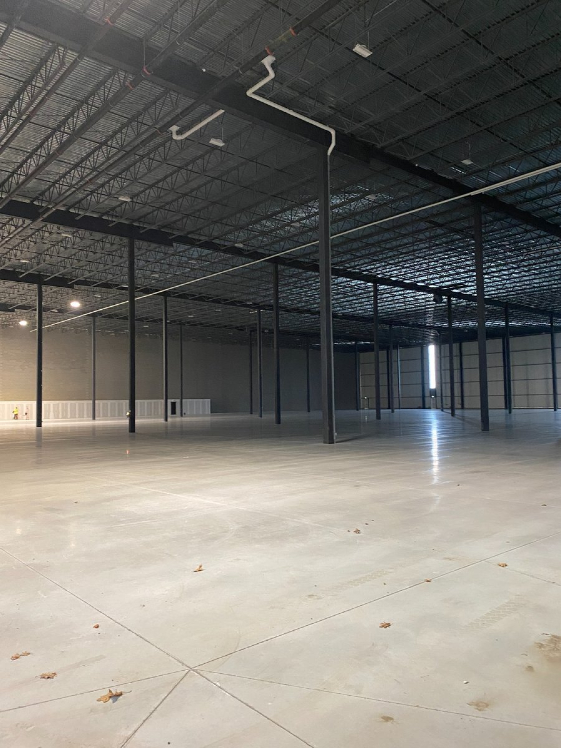 A large, empty industrial warehouse interior with a polished concrete floor, tall steel columns, and exposed ceiling structure. Overhead lighting and visible ductwork span the open space, with perimeter walls in neutral tones.