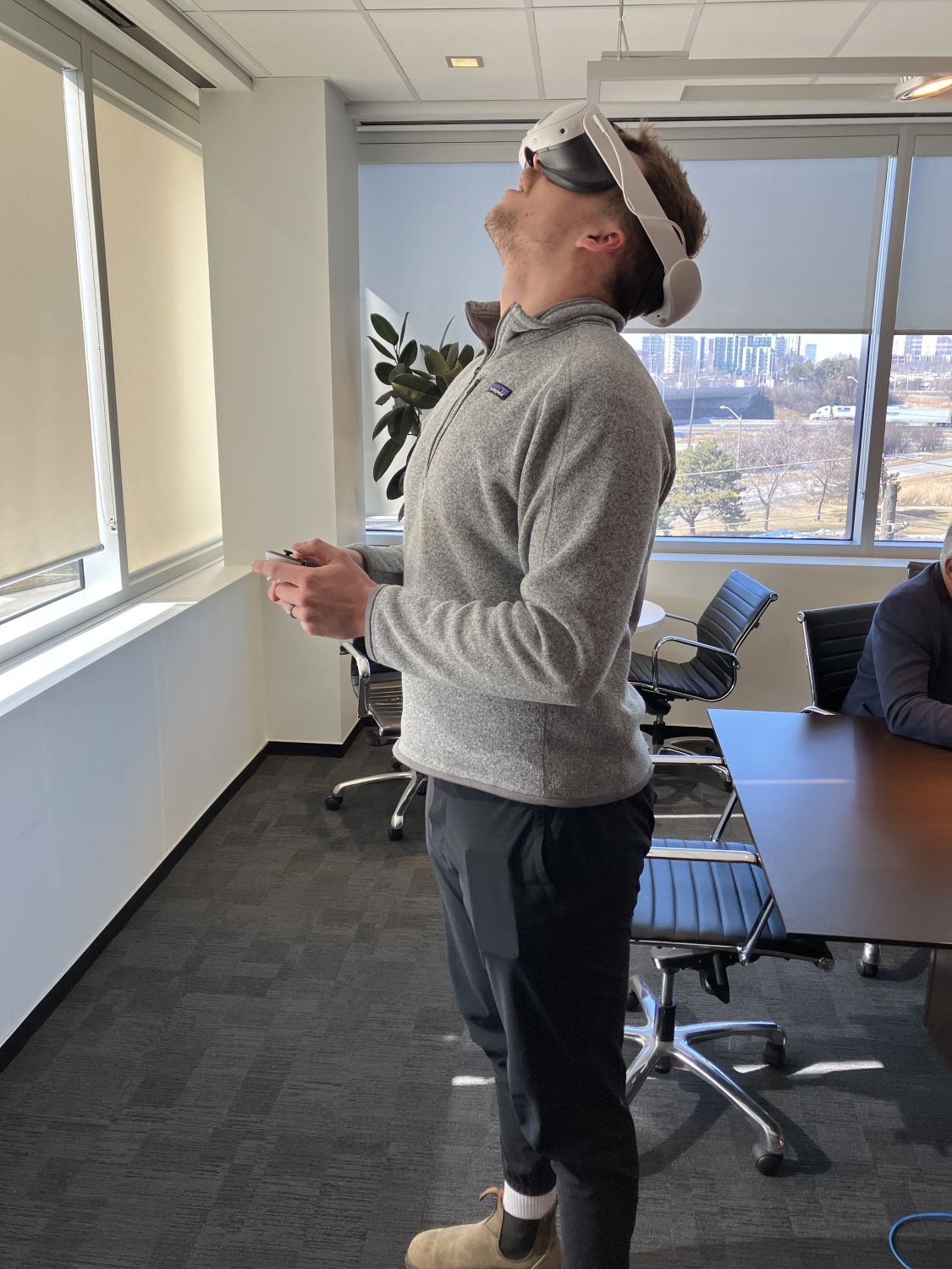An office meeting room with tables and chairs, where a person stands wearing a virtual reality headset and holding controllers. Large windows, blinds, and a city view are visible in the background.