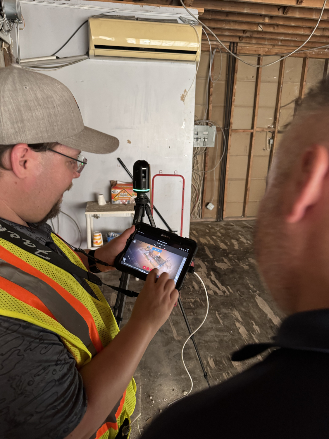 A person wearing safety gear stands inside an unfinished room, operating a tablet connected to a tripod‑mounted 3D scanning device. Exposed studs, wiring, and flooring indicate an interior space under renovation.