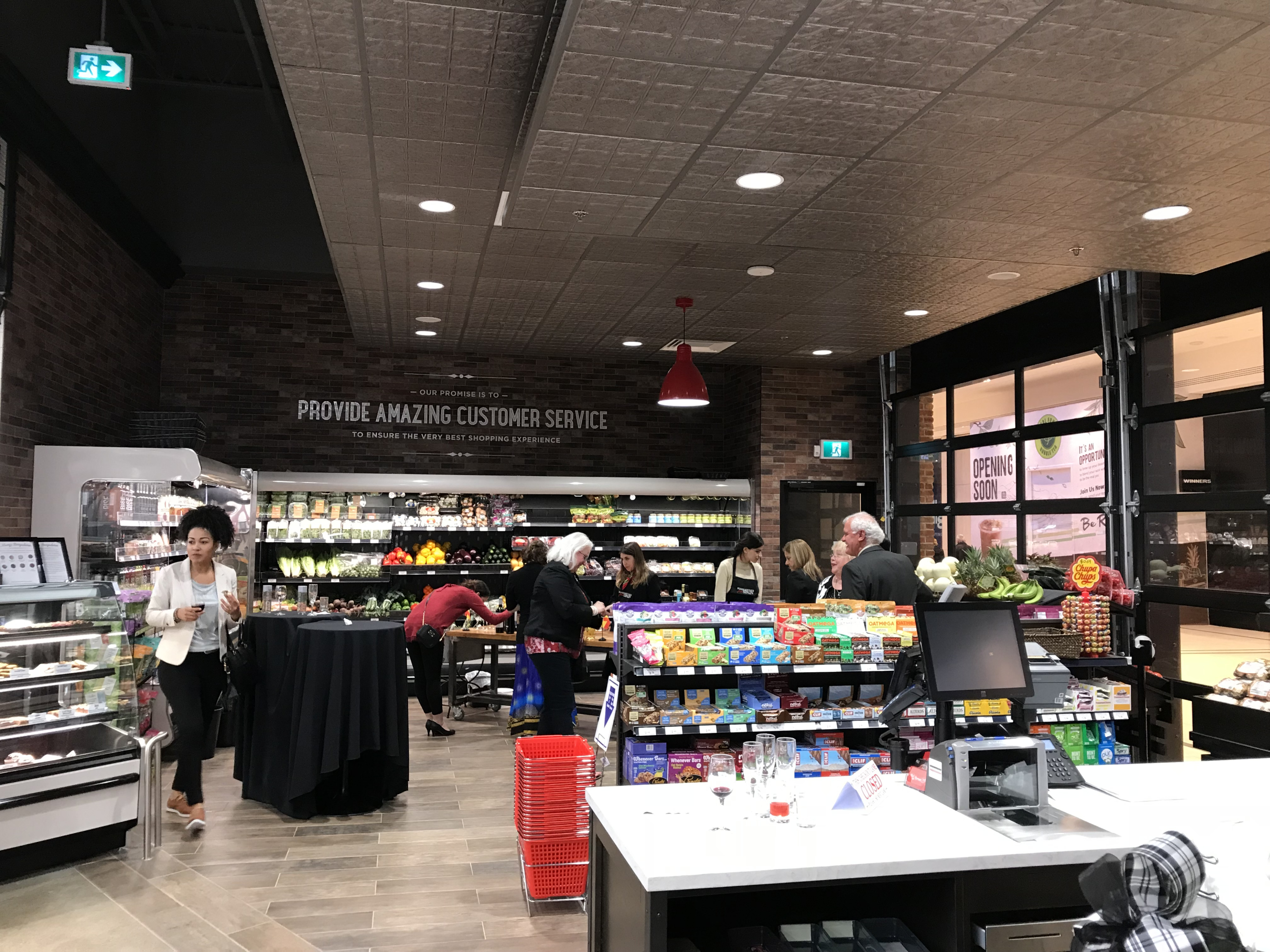 A grocery store interior with checkout counters, refrigerated displays, and shelves of packaged goods. Shoppers browse and line up at the tills beneath red signage and bright overhead lighting.
