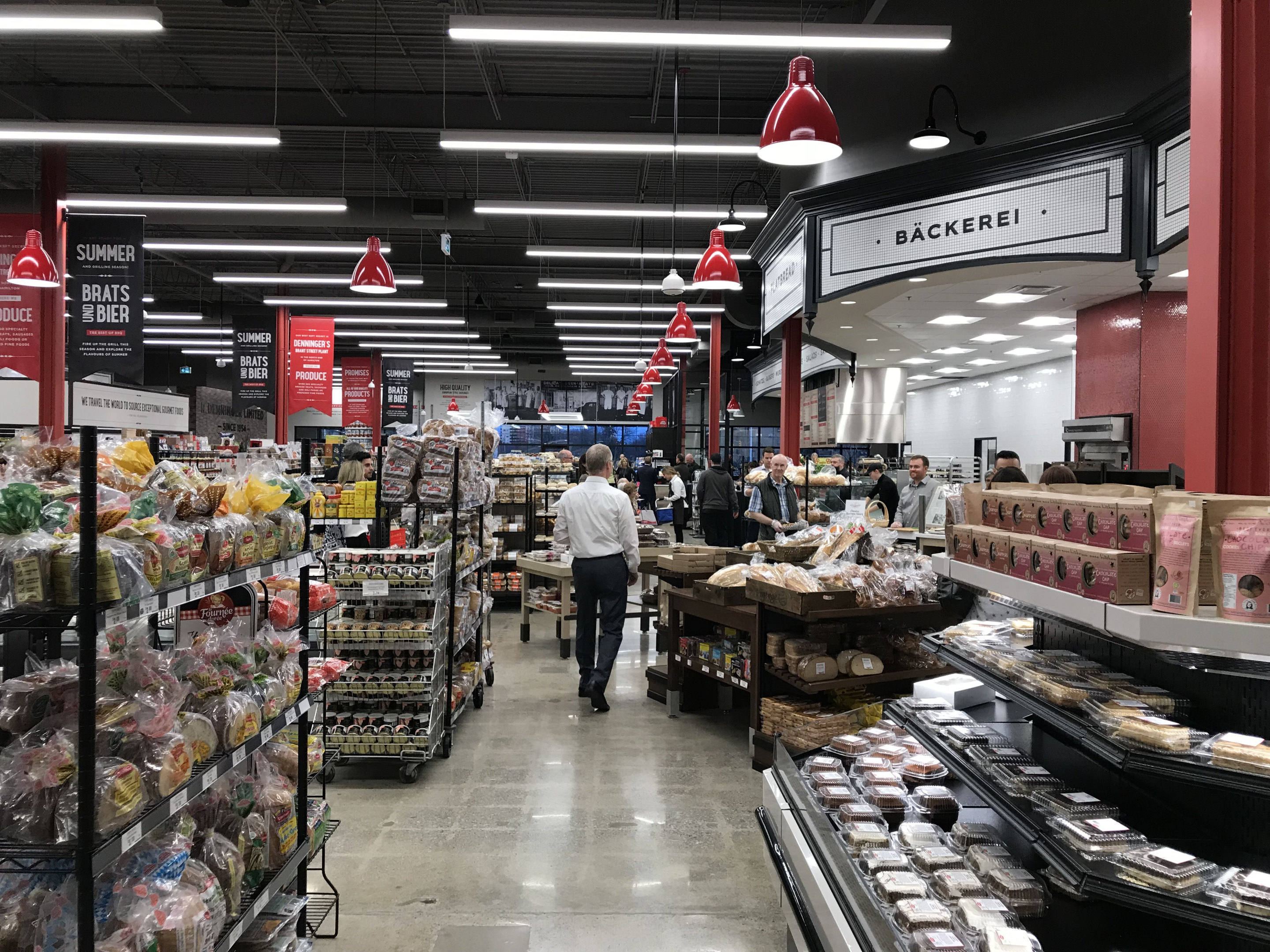 A grocery store bakery aisle with shelves and refrigerated cases displaying packaged breads and pastries. Shoppers move through the wide aisle under bright linear lighting and red hanging signs.