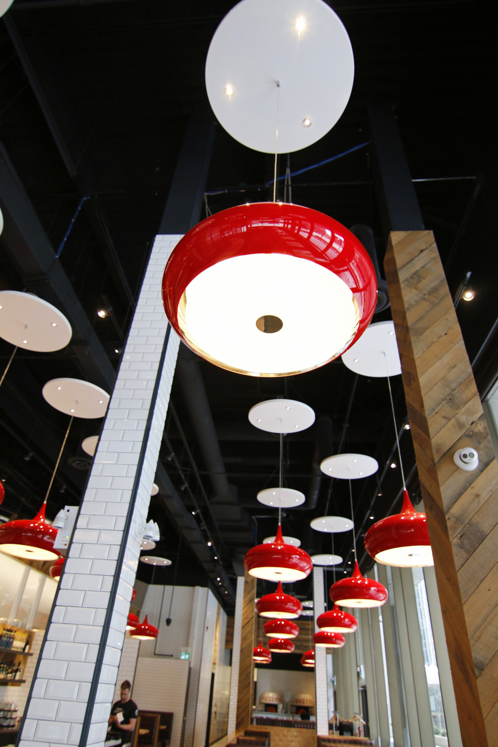 A restaurant interior ceiling with rows of red and white circular pendant lights hanging at varying heights. The lights run along a long dining space with black ceilings, wood accents, and white tiled columns.